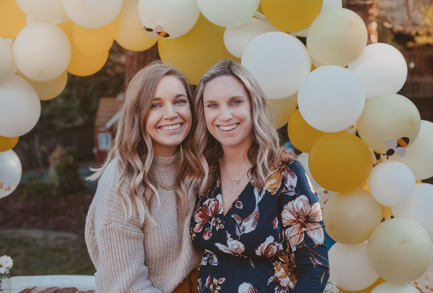 Two smiling women pose together under a yellow and white balloon arch at an outdoor celebration.