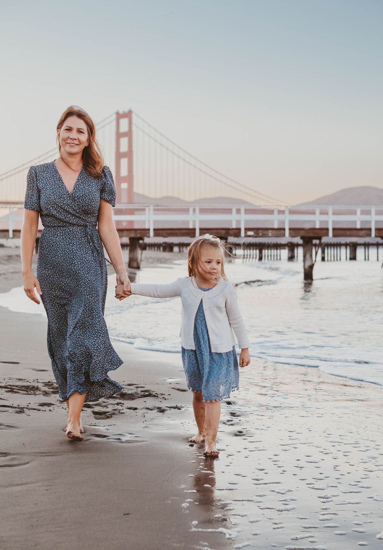 Mother and young daughter walking hand-in-hand along the beach near the Golden Gate Bridge.