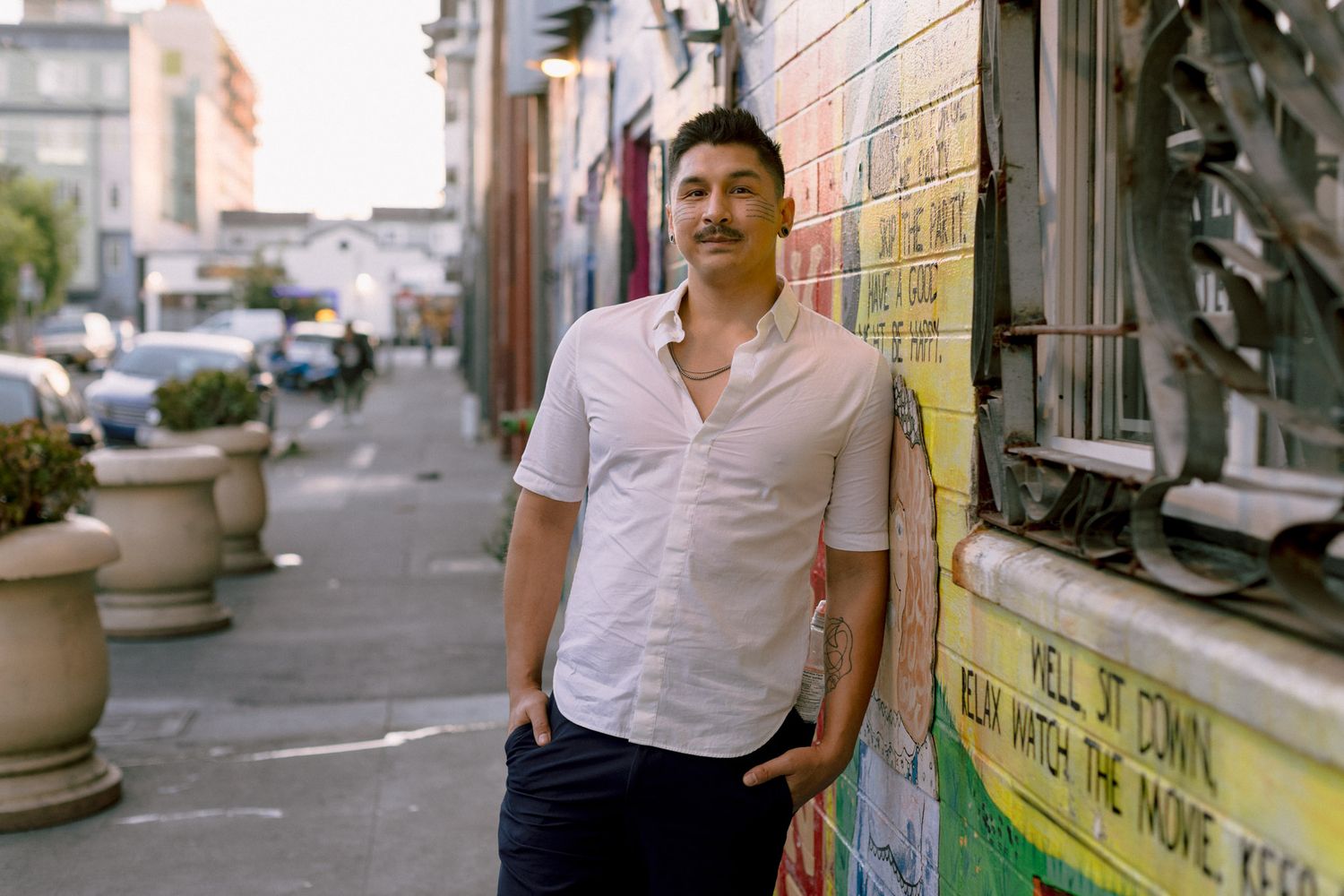 Man in white shirt leaning against colorful mural wall on urban street with potted plants.