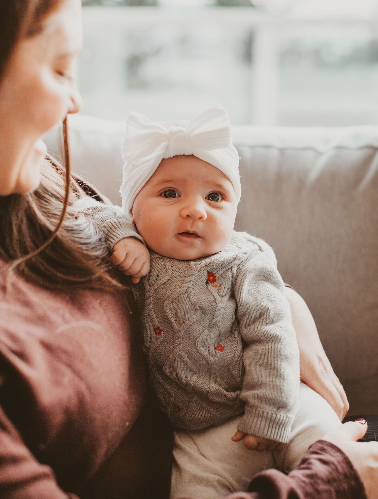 Cute baby girl in gray knit sweater and white bow headband being held by mother on couch.