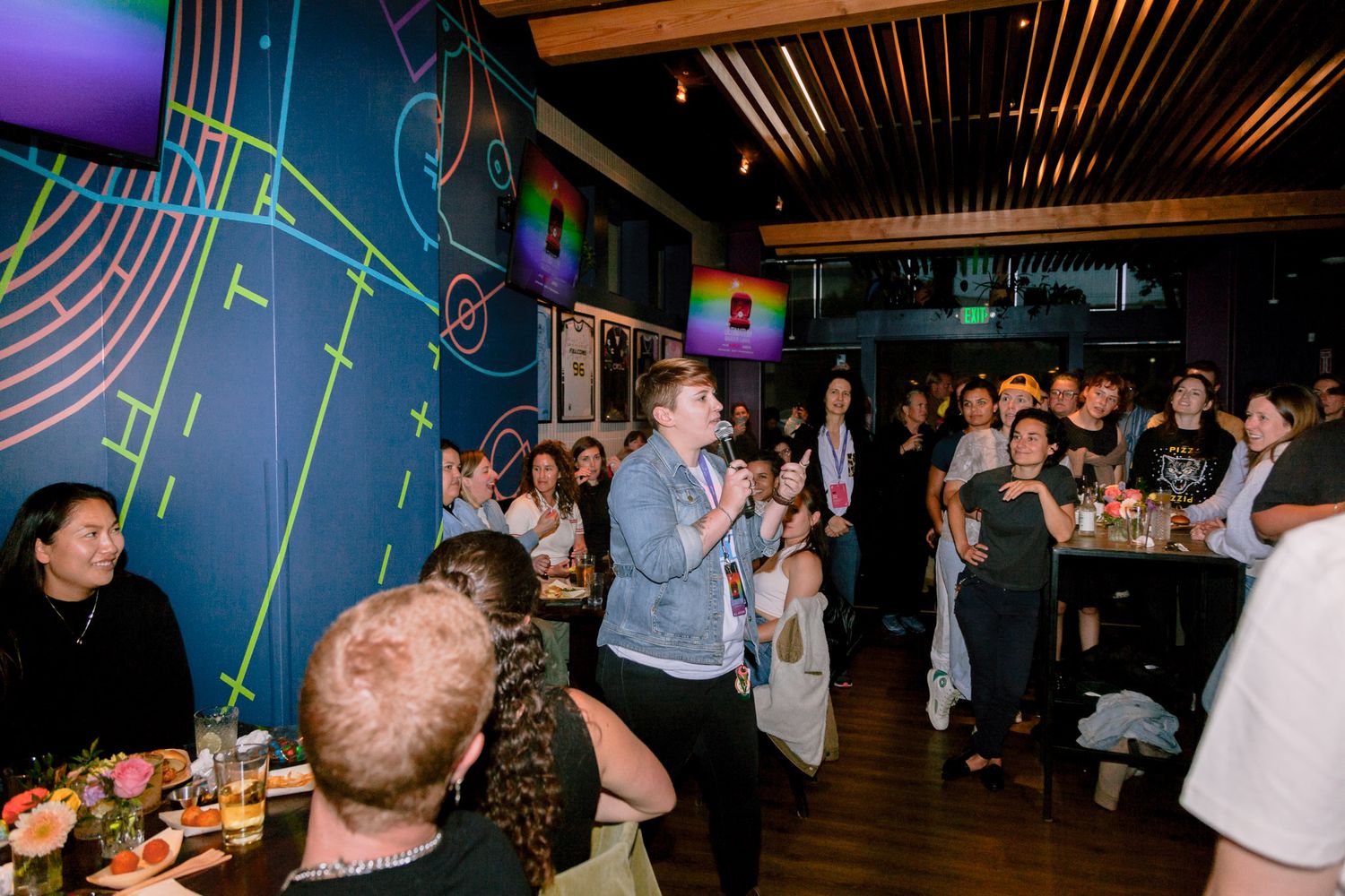 A performer speaks into a microphone at a lively indoor event with colorful neon decor and an engaged crowd.