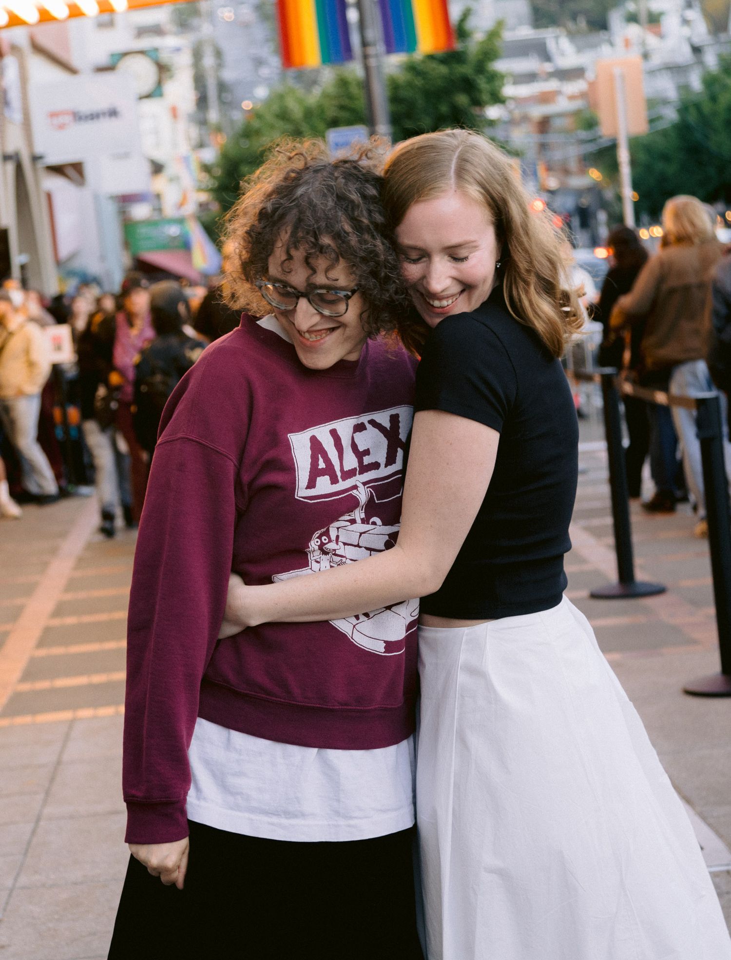 Two young women hugging and smiling on a busy street with rainbow pride flags in background.