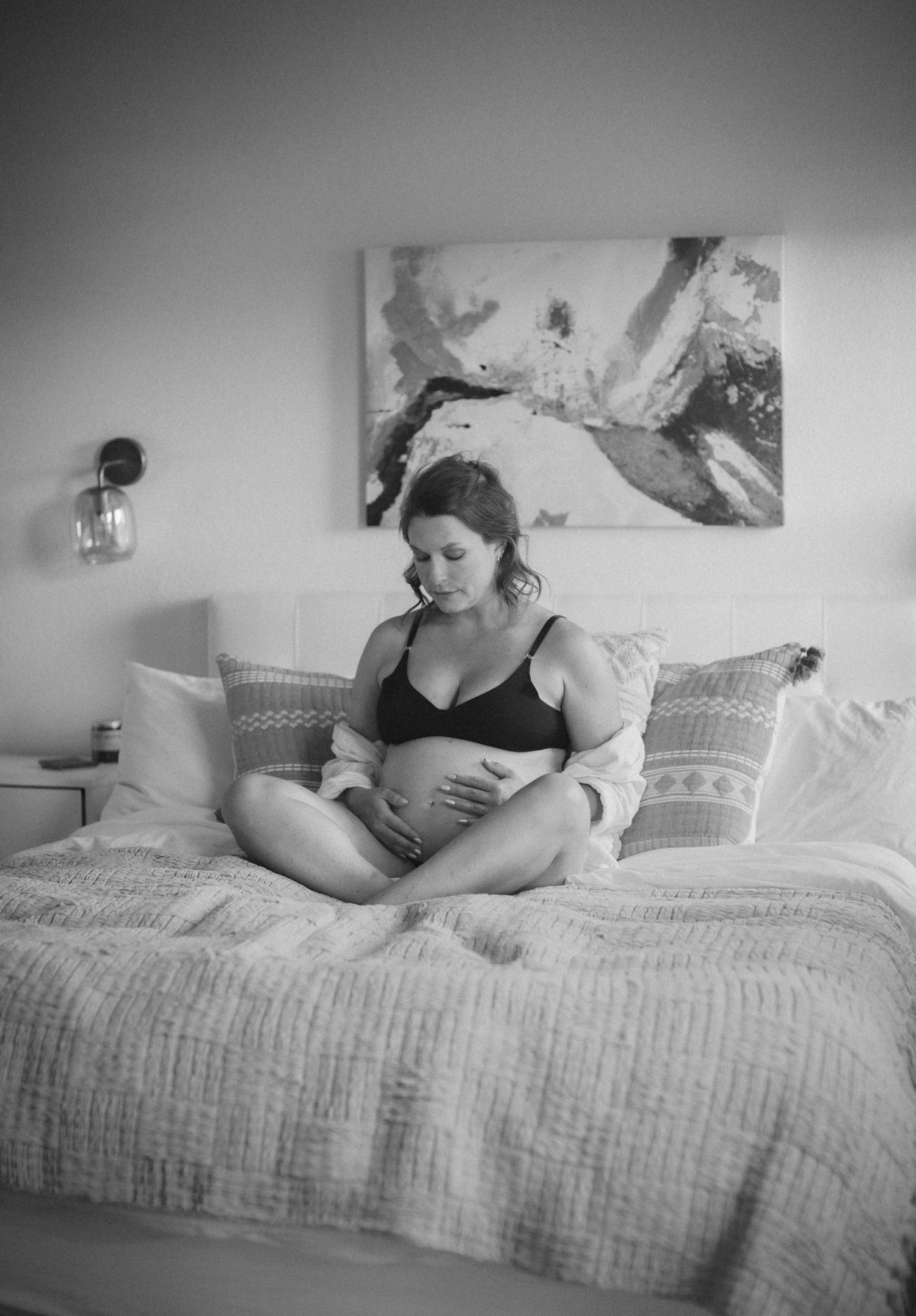 Black and white photo of pregnant woman sitting on bed cradling her baby bump.