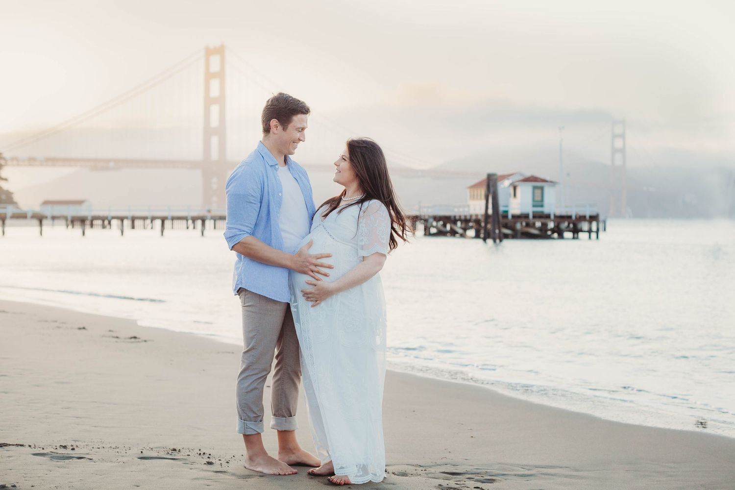 Pregnant couple embracing on beach with Golden Gate Bridge in background at sunset.