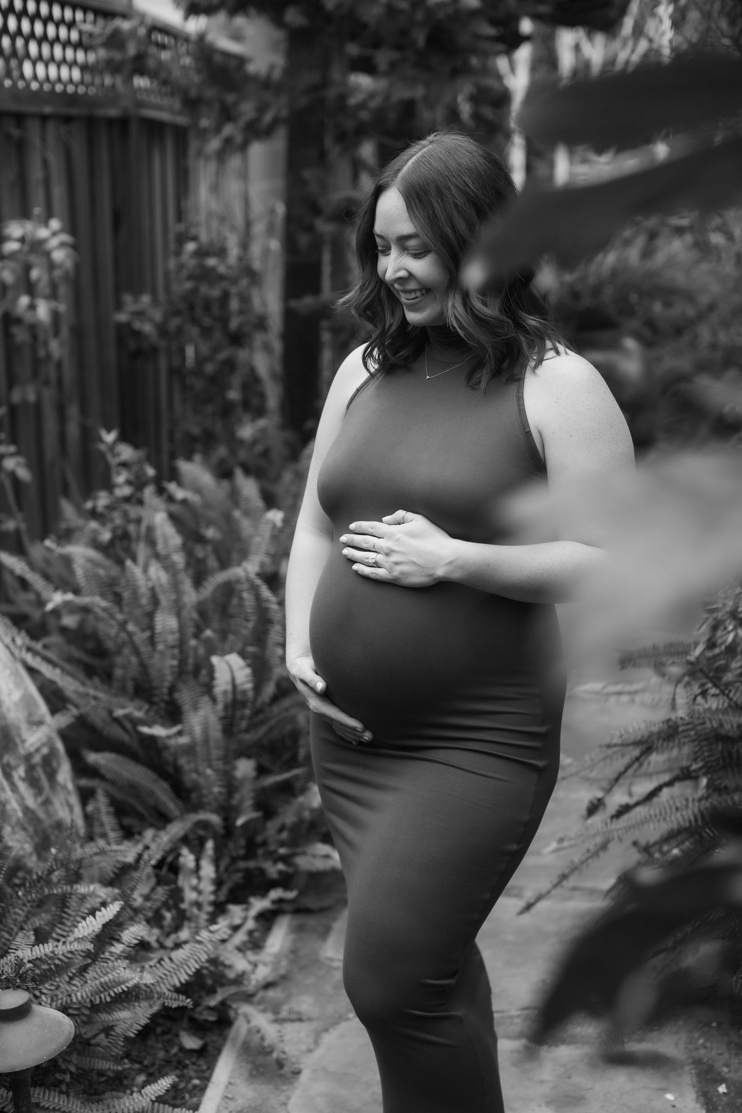 Black and white photo of a smiling pregnant woman in a fitted dress cradling her baby bump outdoors among lush greenery.