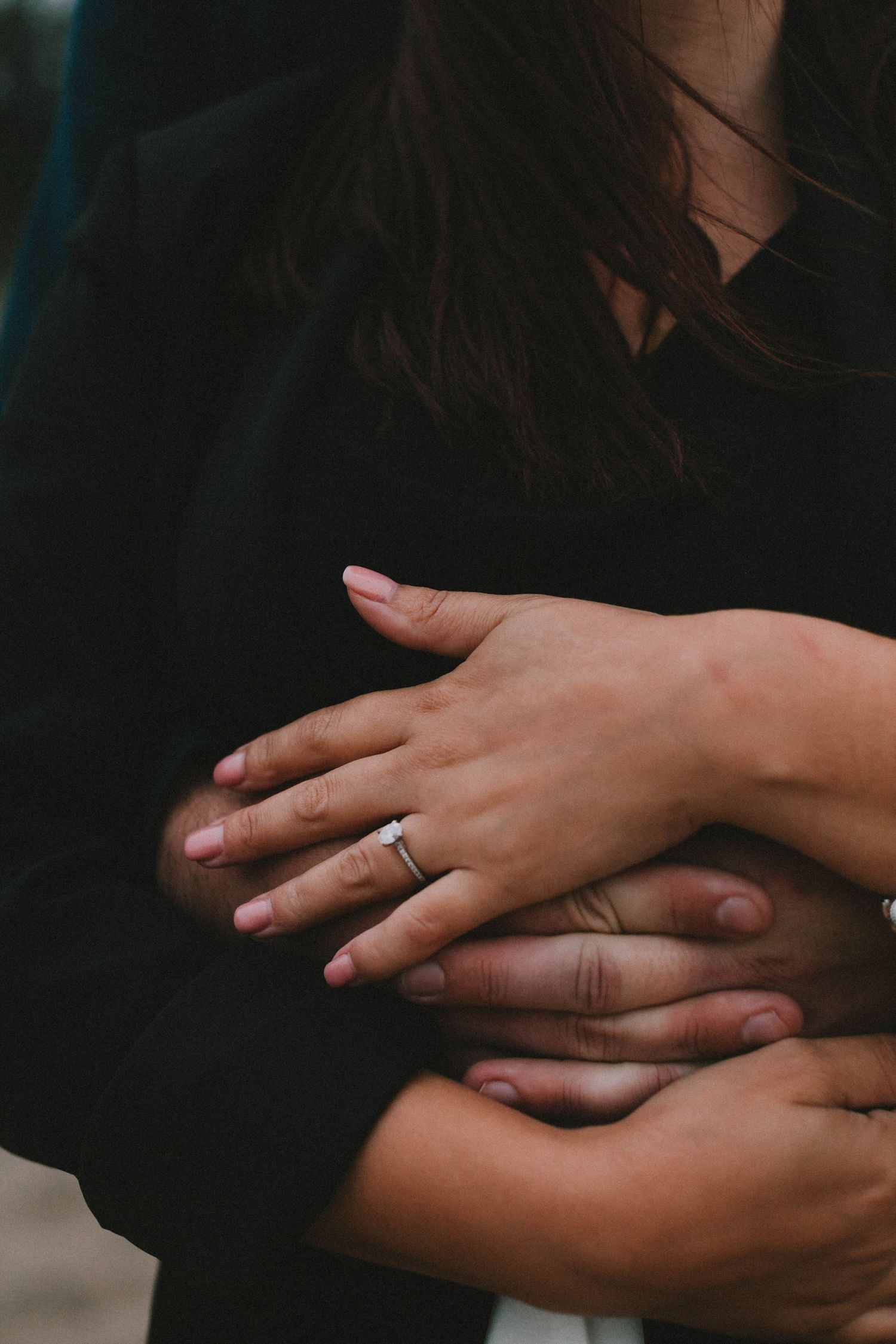 Close-up of hands embracing, woman wearing a diamond engagement ring, couple in dark clothing.