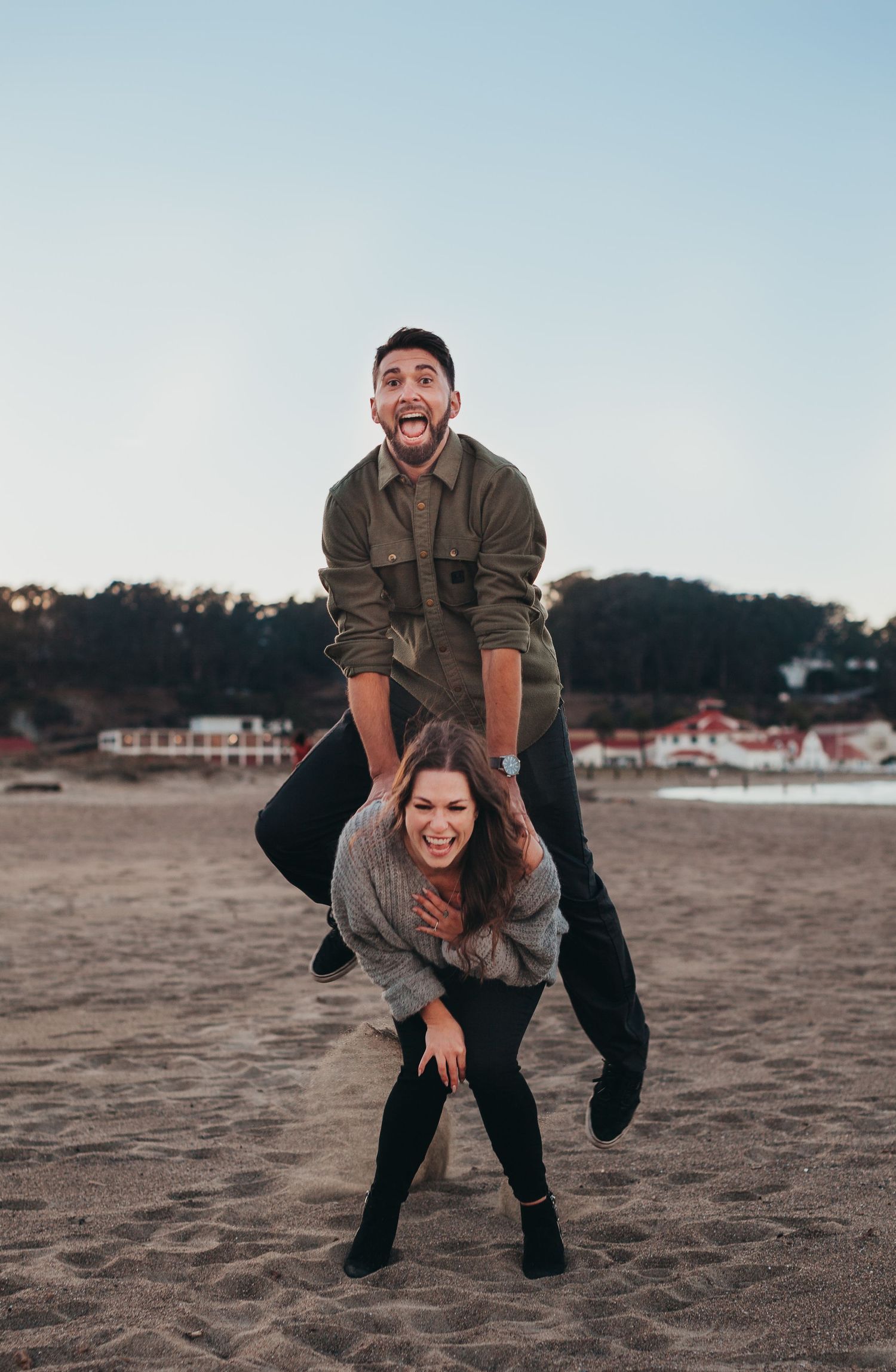 Playful couple on beach, playing leap frog, both laughing.