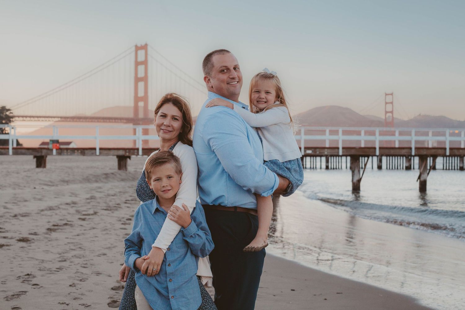 Happy family of four posing on Baker Beach with the Golden Gate Bridge at sunset.