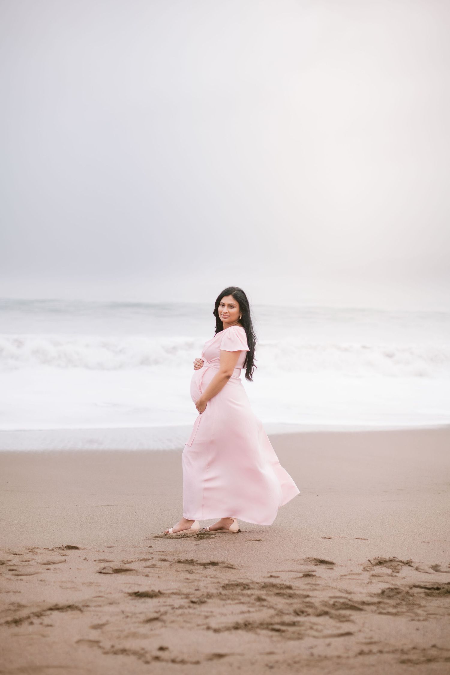 Pregnant woman in pink flowing dress standing on beach with ocean waves behind her.