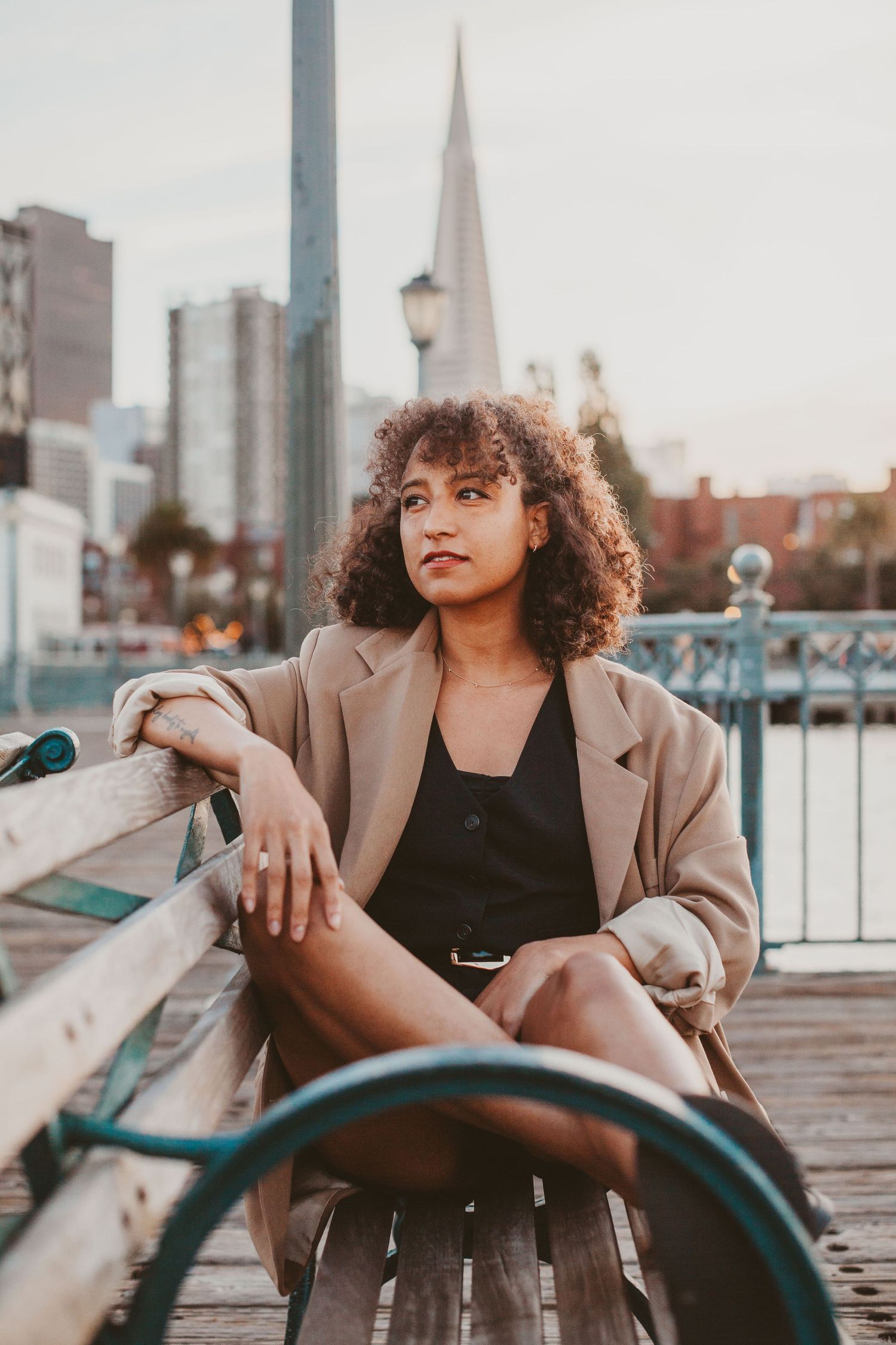 Young woman in beige blazer sitting on wooden bench with San Francisco skyline and Transamerica Pyramid in background.