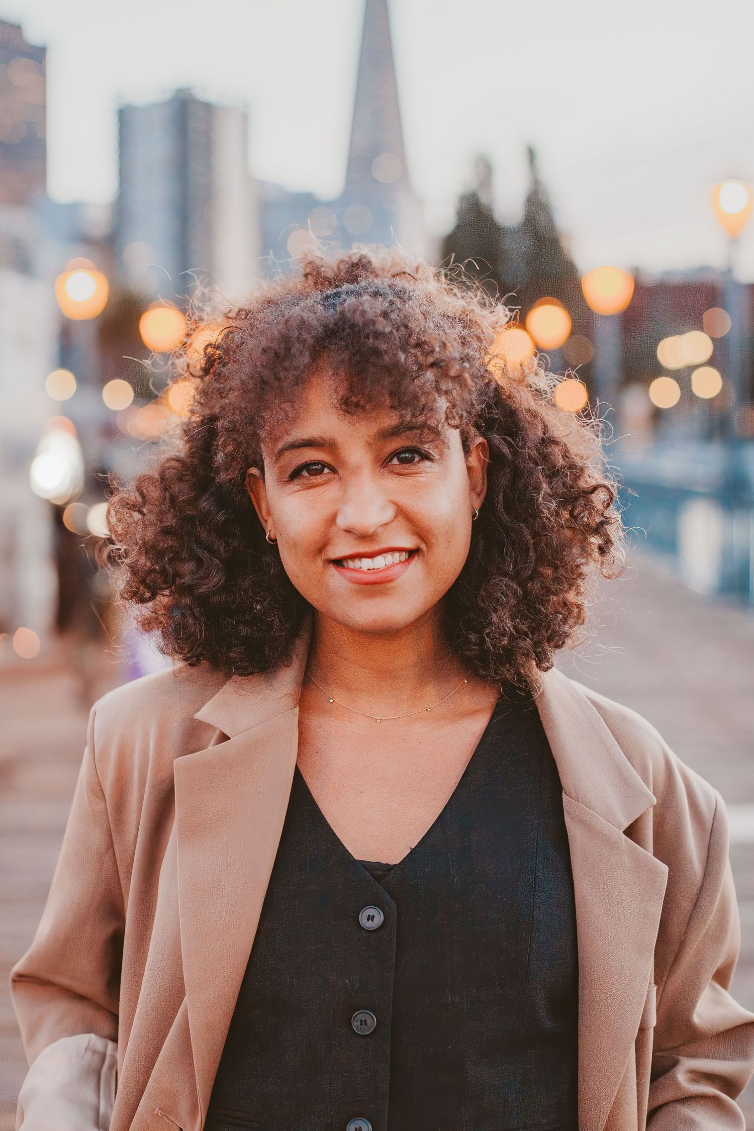 Smiling young woman with curly hair wearing a beige blazer outdoors with bokeh city lights at dusk.