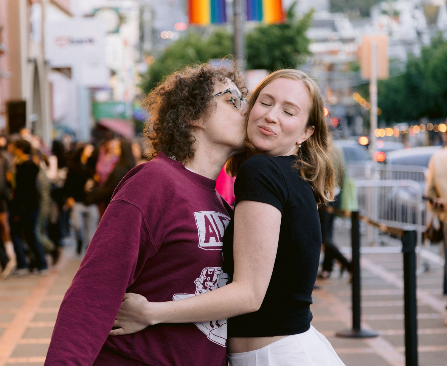 Two people share a kiss on a city street decorated with rainbow Pride flags at dusk.