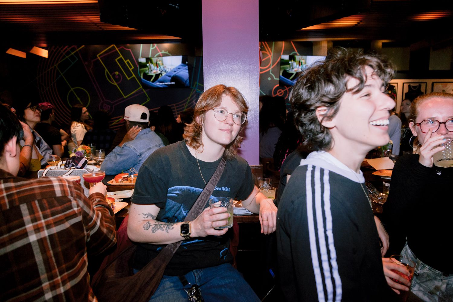 Two young people smile and hold drinks at a casual indoor social gathering with friends in the background.