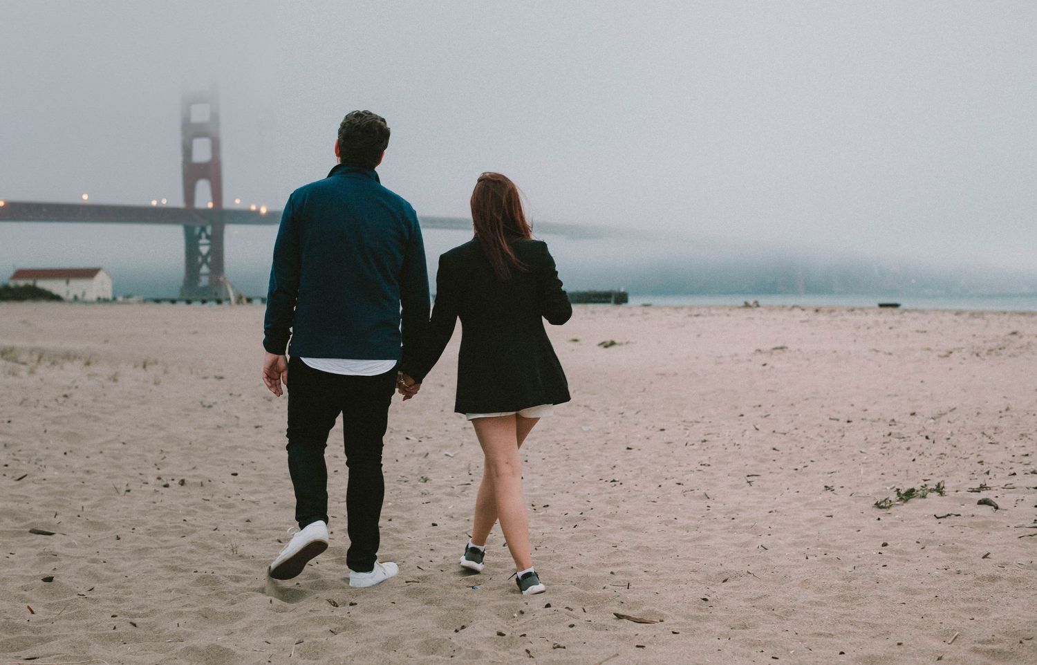 Couple holding hands walking on a sandy beach near the foggy Golden Gate Bridge.