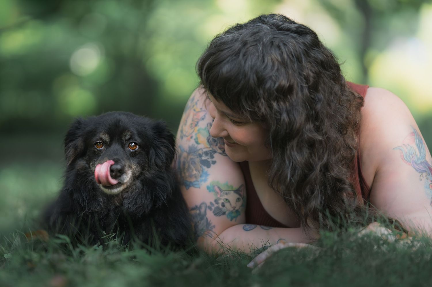 Tattooed woman lying on grass smiling at her black fluffy dog licking its nose in a green park.