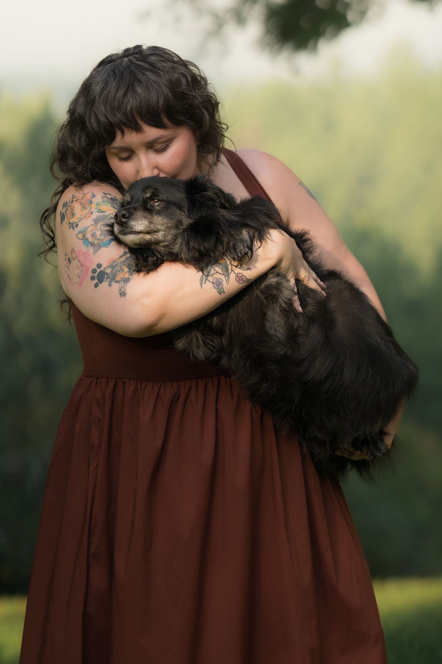 Woman with tattoos in burgundy dress lovingly hugging a small black dog outdoors with green trees behind.