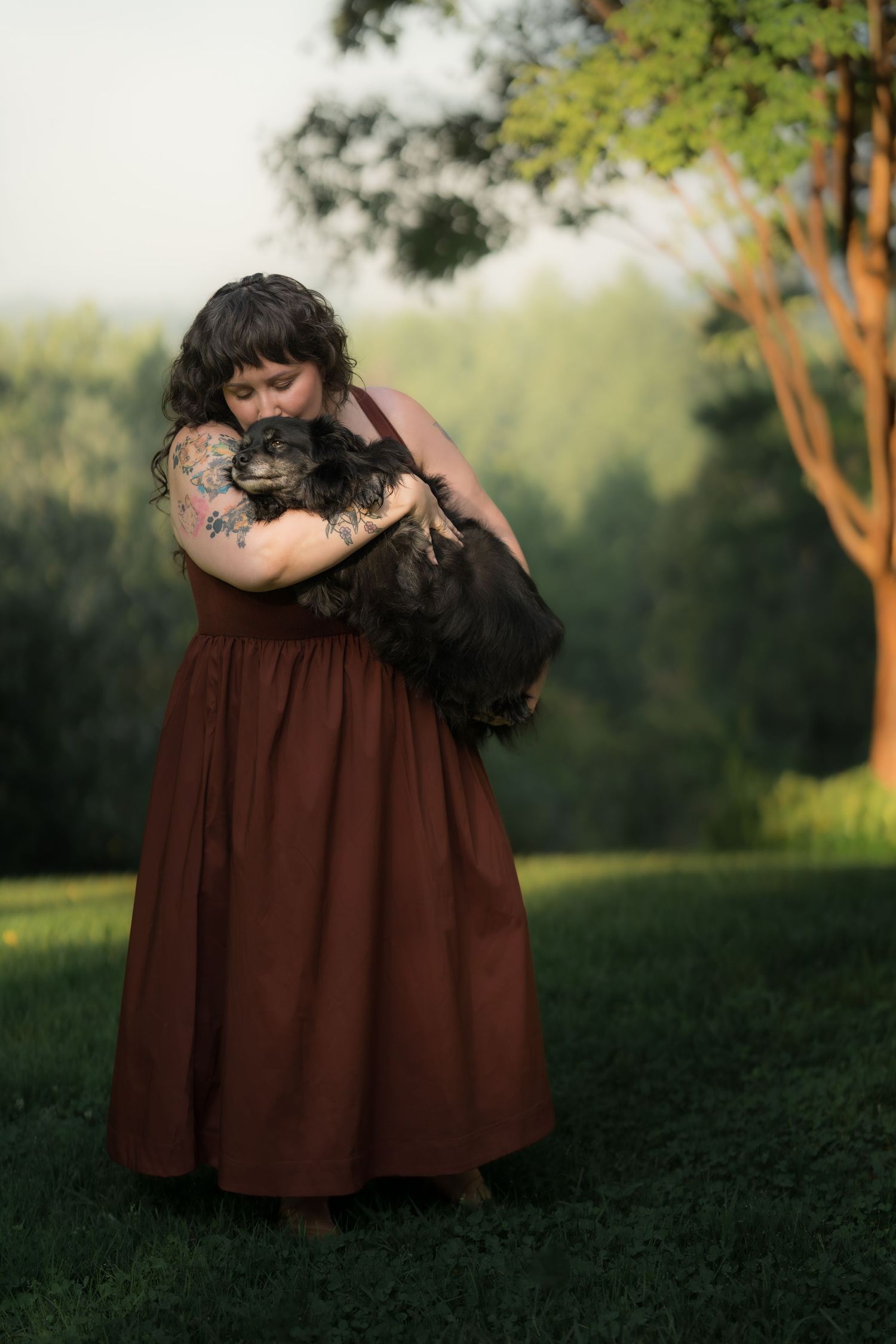 Woman in rust dress hugging a small black dog outdoors in a lush green park setting.