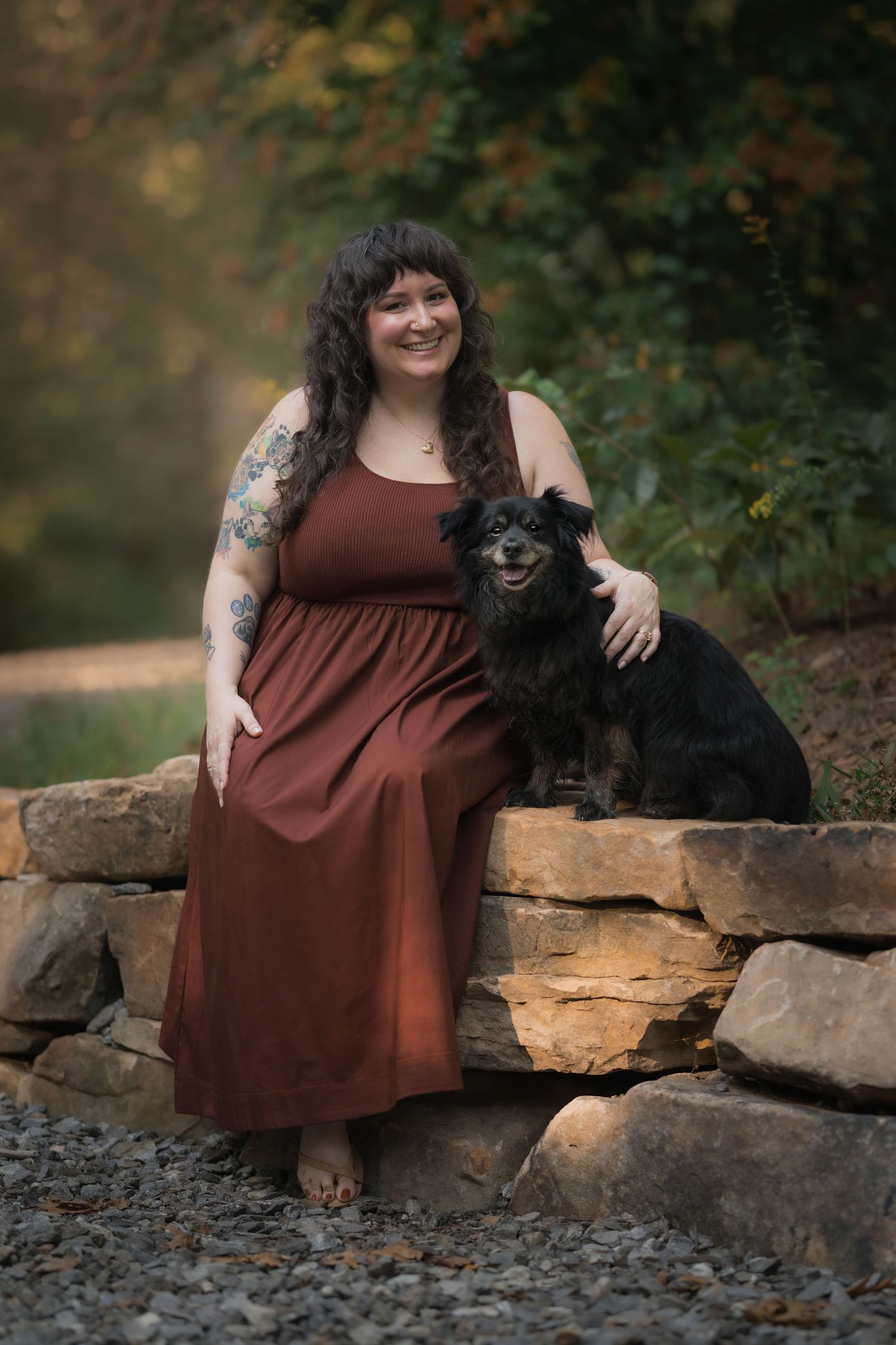 A smiling woman in a rust maxi dress sits on a stone wall outdoors with a small black dog beside her.