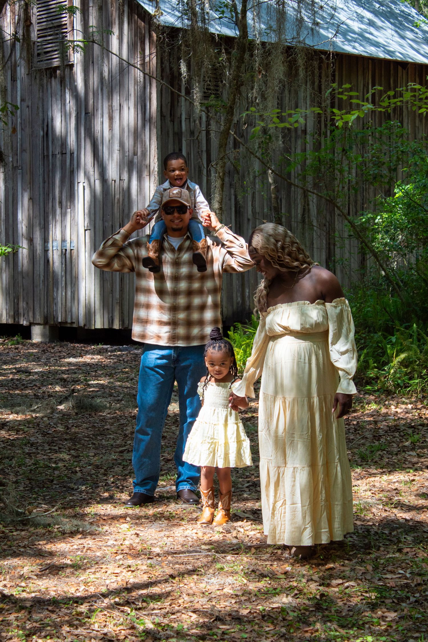 Pregnant Black woman and man with child on shoulders stand near rustic barn outdoors.