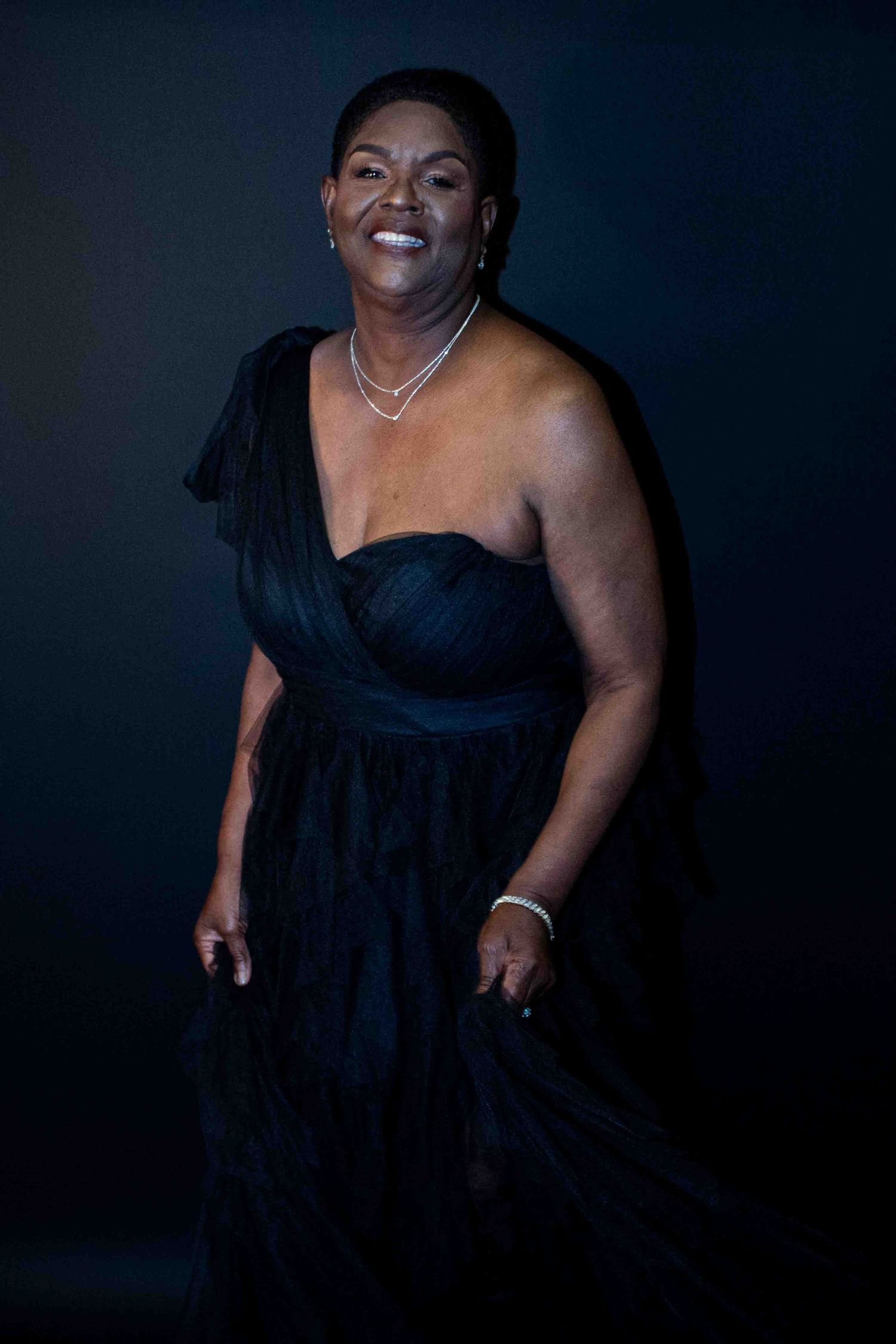 Woman in elegant black one-shoulder gown smiling confidently against dark background.