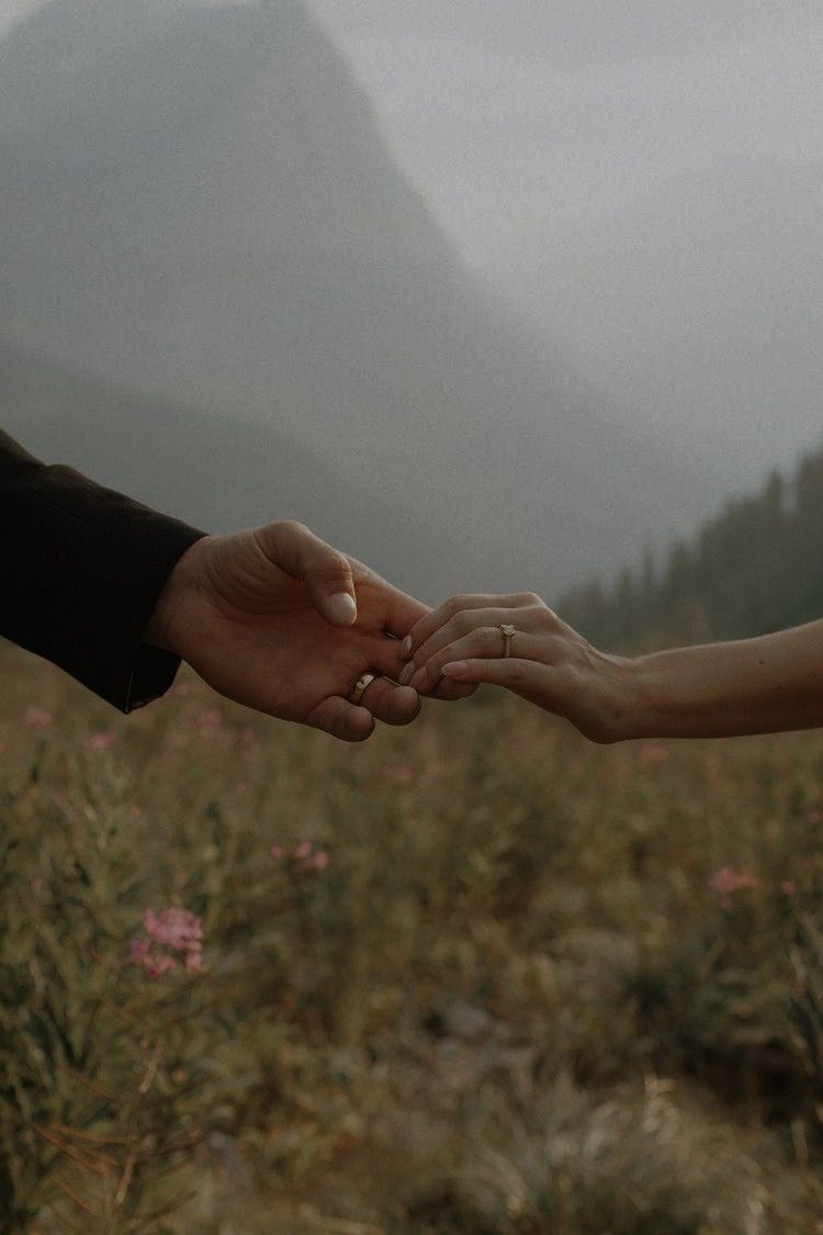 Close-up of two people gently holding hands in a misty mountain meadow with wildflowers in background.