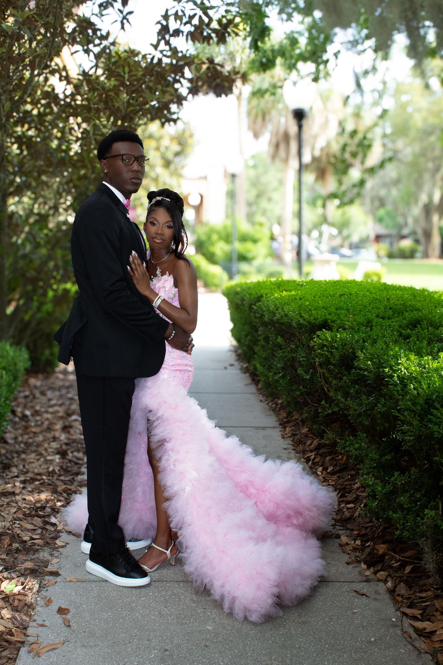Young Black couple in formal prom attire pose on sidewalk lined with green hedges.