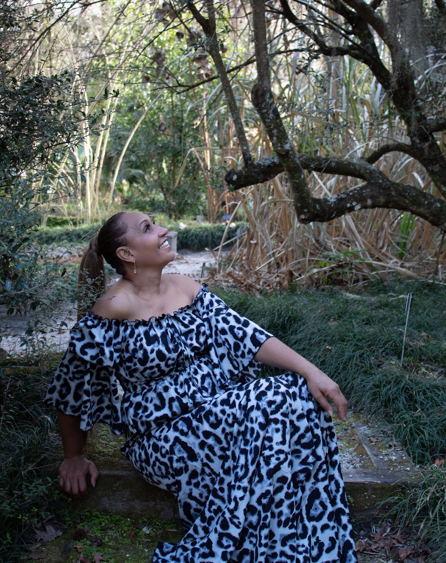 Woman in black and white leopard print off-shoulder dress sitting on steps in a wooded garden.