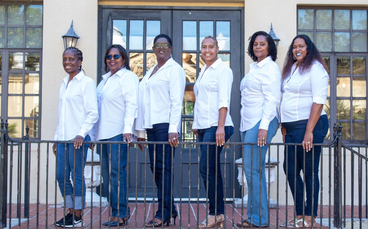 Six women in matching white shirts and jeans posing in a line in front of a building.
