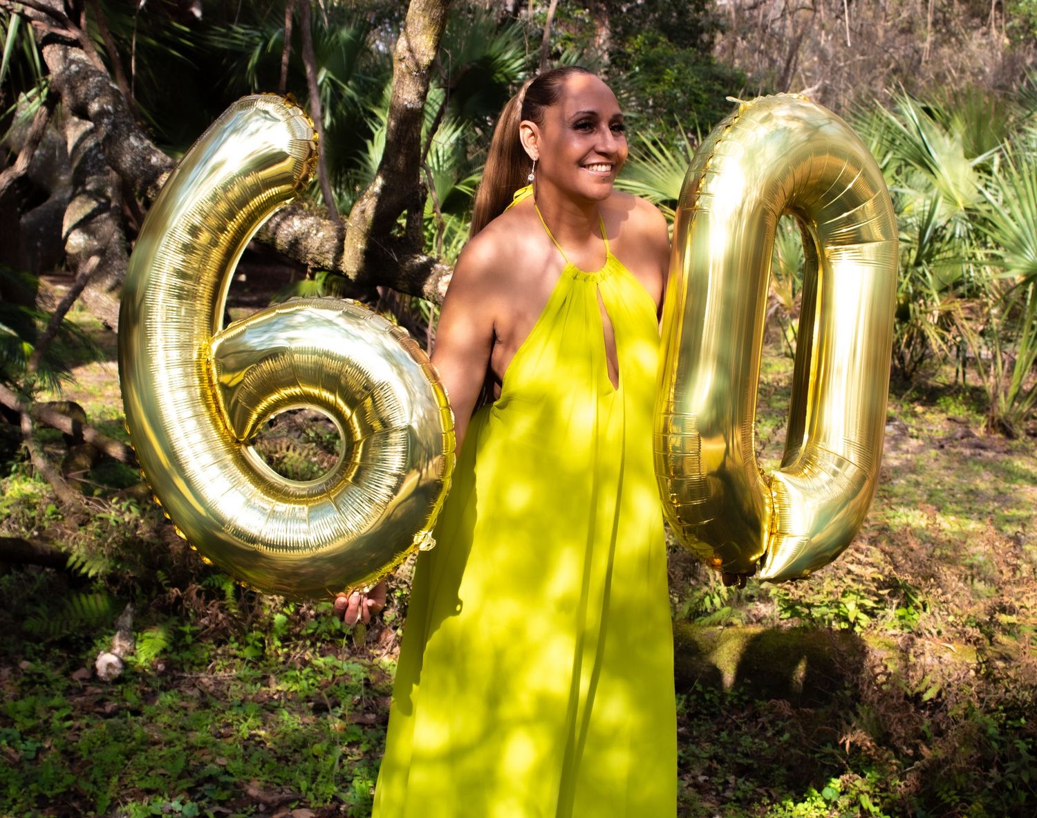 Woman in yellow dress celebrating 60th birthday with gold number balloons in a tropical garden.