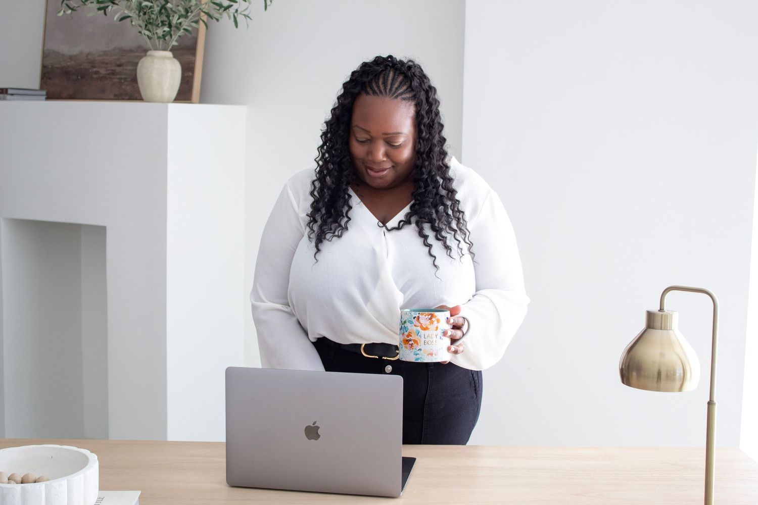 Black woman in white blouse holding mug while working on MacBook laptop at desk.