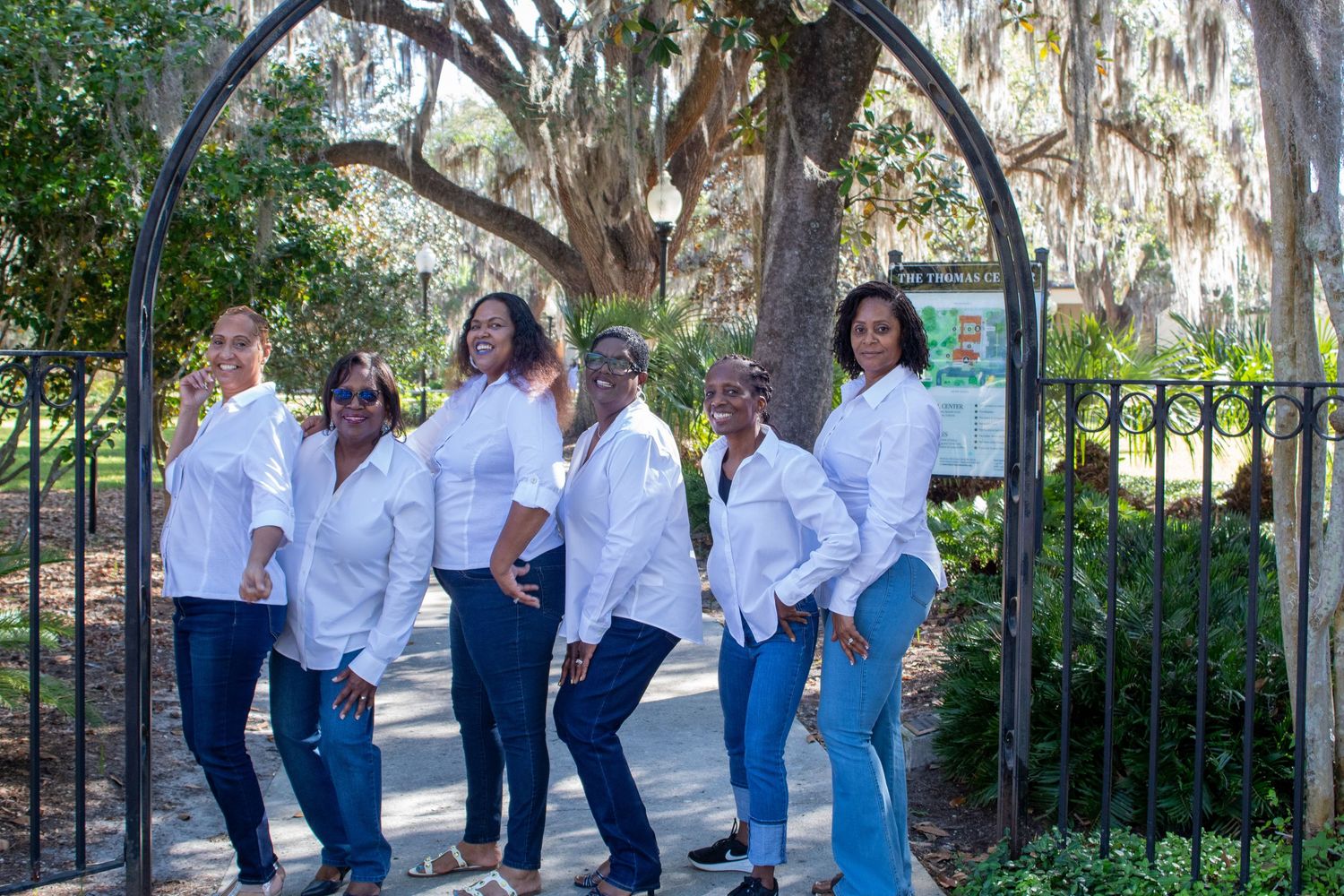 Six women in white shirts and jeans posing under an iron arch surrounded by Spanish moss trees.