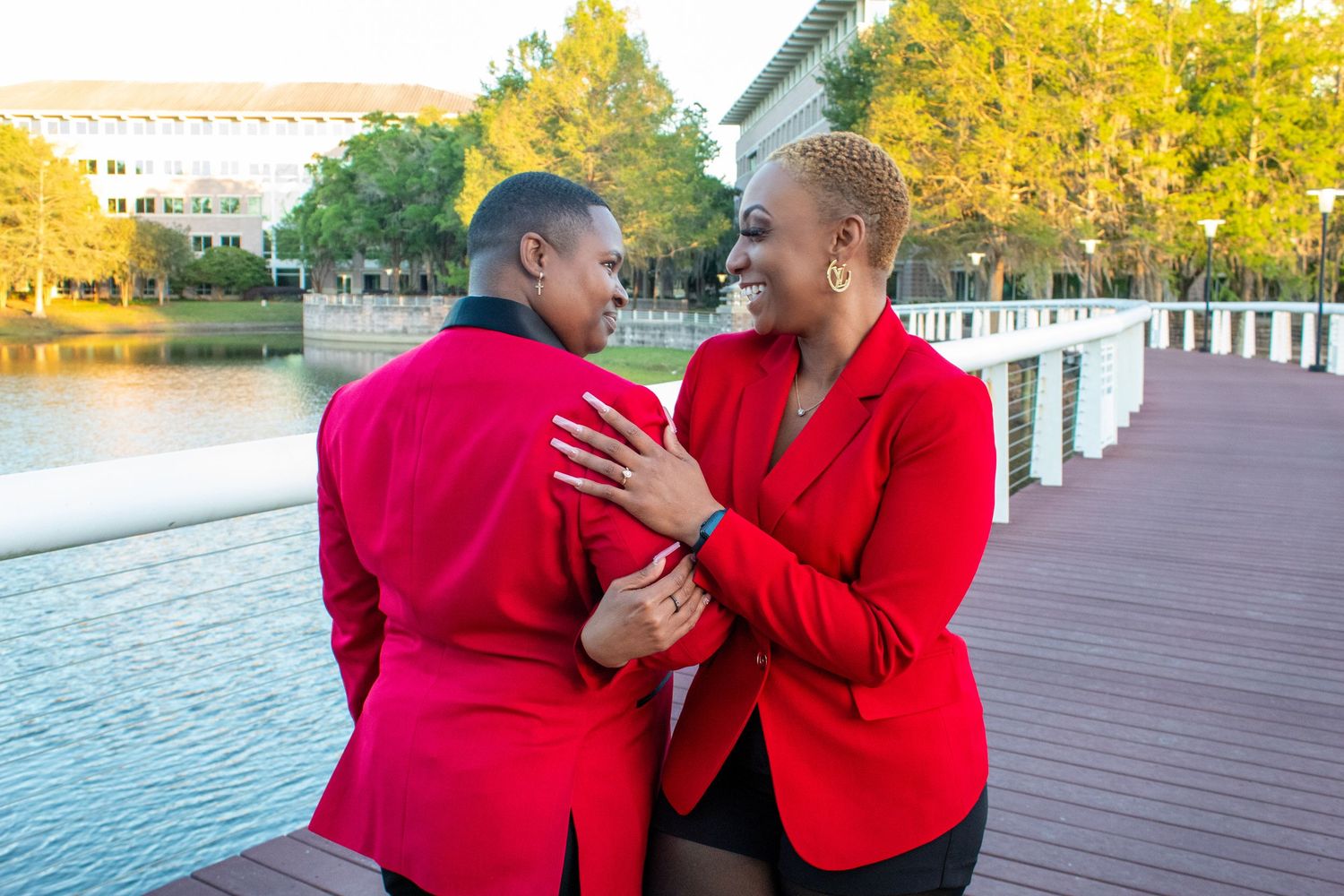 Two women in matching red blazers embracing and smiling near a waterfront boardwalk at sunset.