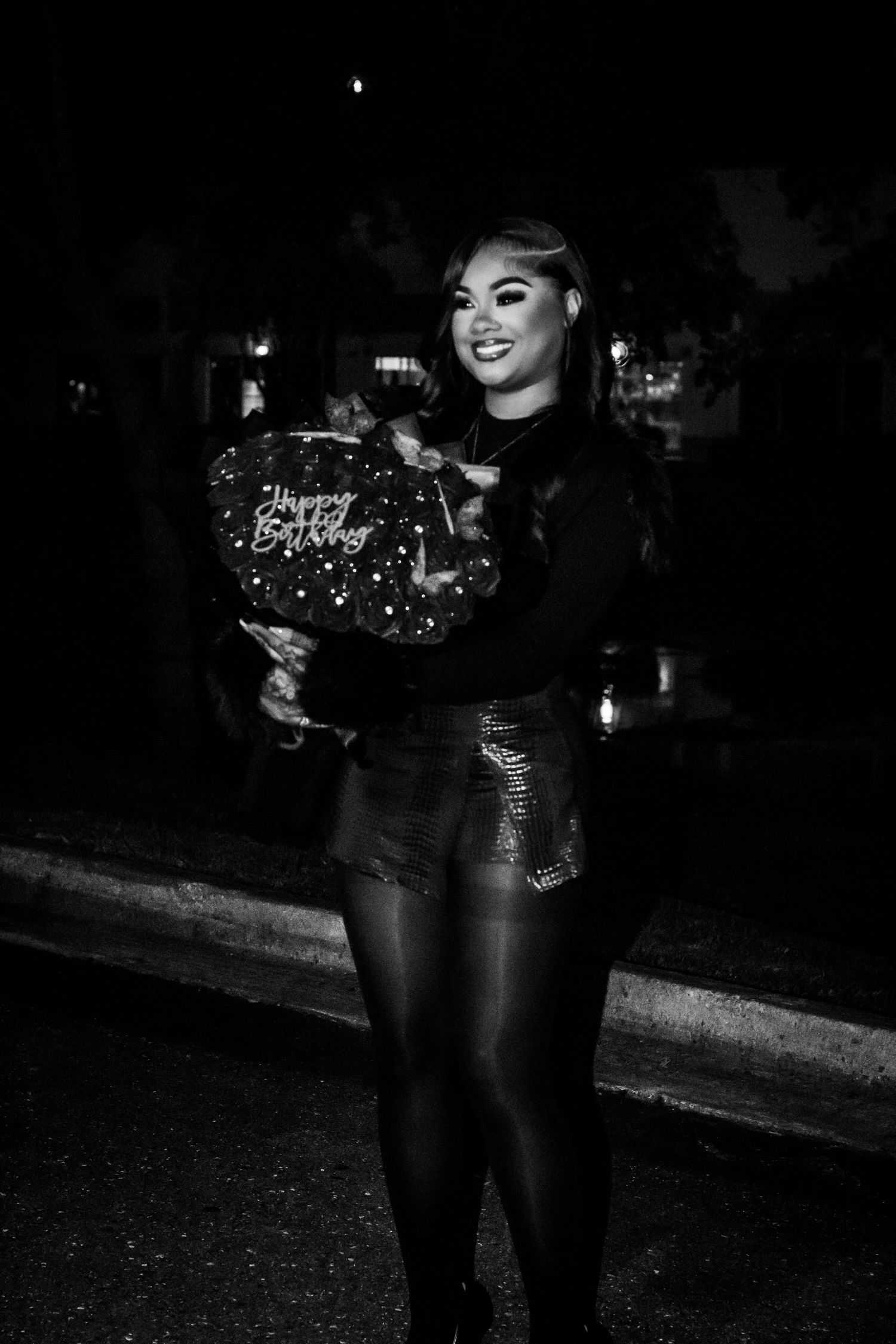 Black and white photo of woman holding Happy Birthday balloon bouquet outdoors at night.