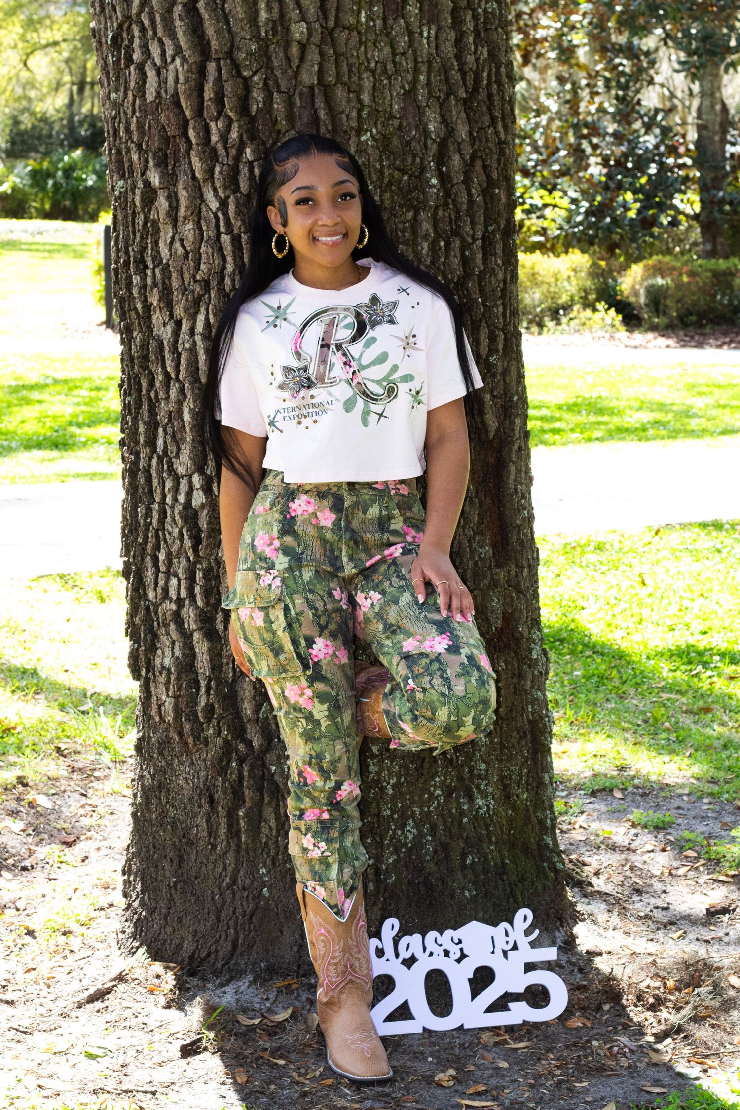 High school senior girl posing by a tree with Class of 2025 sign outdoors.