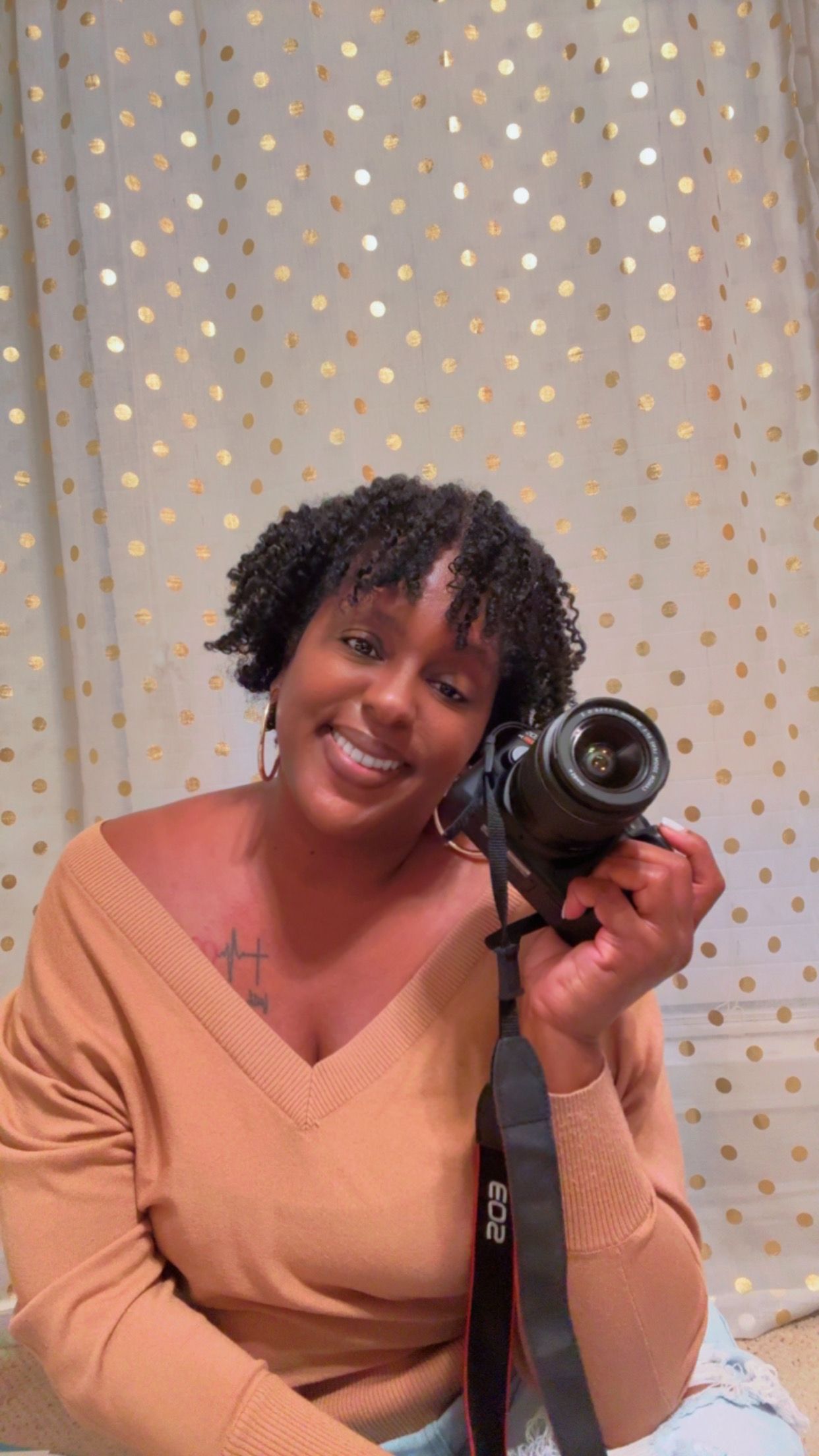 Smiling woman holding Canon DSLR camera in front of gold polka dot backdrop curtain.