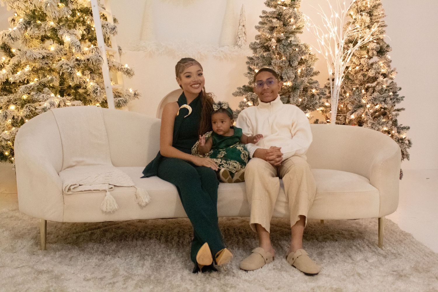 Young couple with baby sitting on white couch in front of Christmas trees.