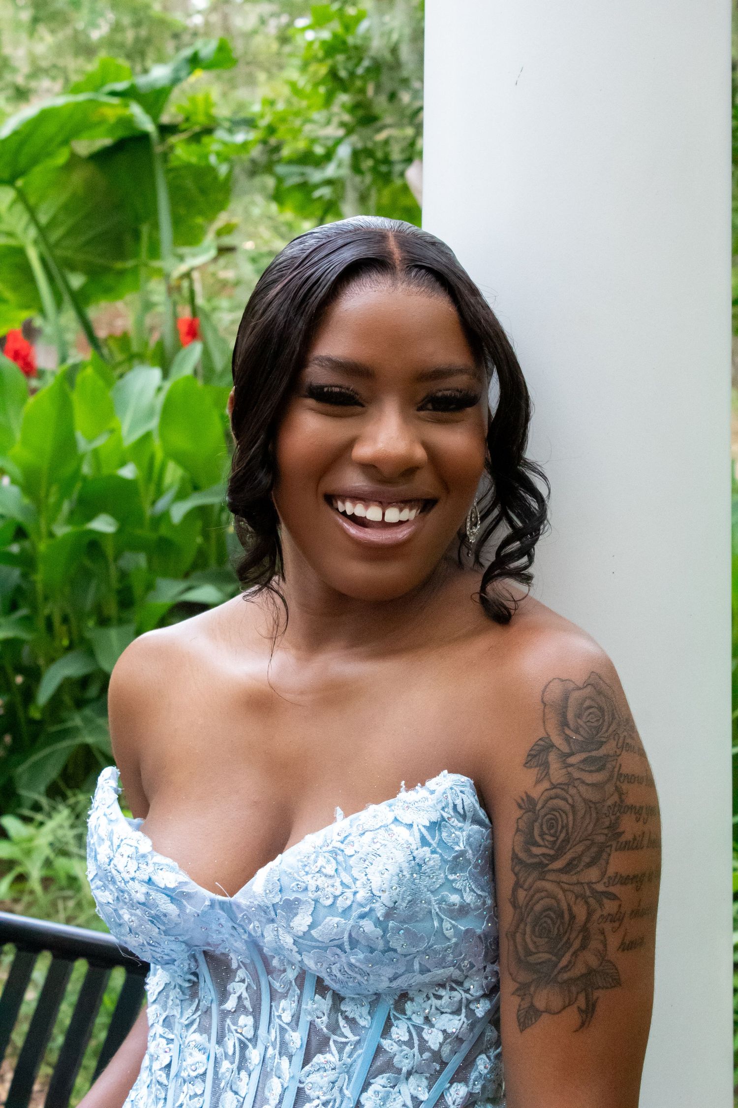 Smiling woman in blue lace strapless gown with rose tattoo on arm leaning against white column outdoors.