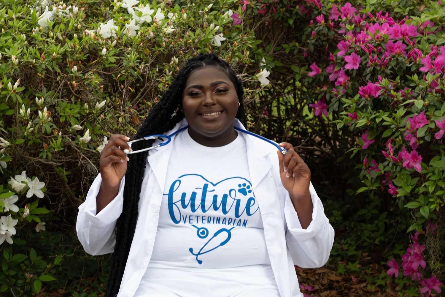 Smiling woman in white lab coat holds stethoscope wearing future veterinarian shirt outdoors.