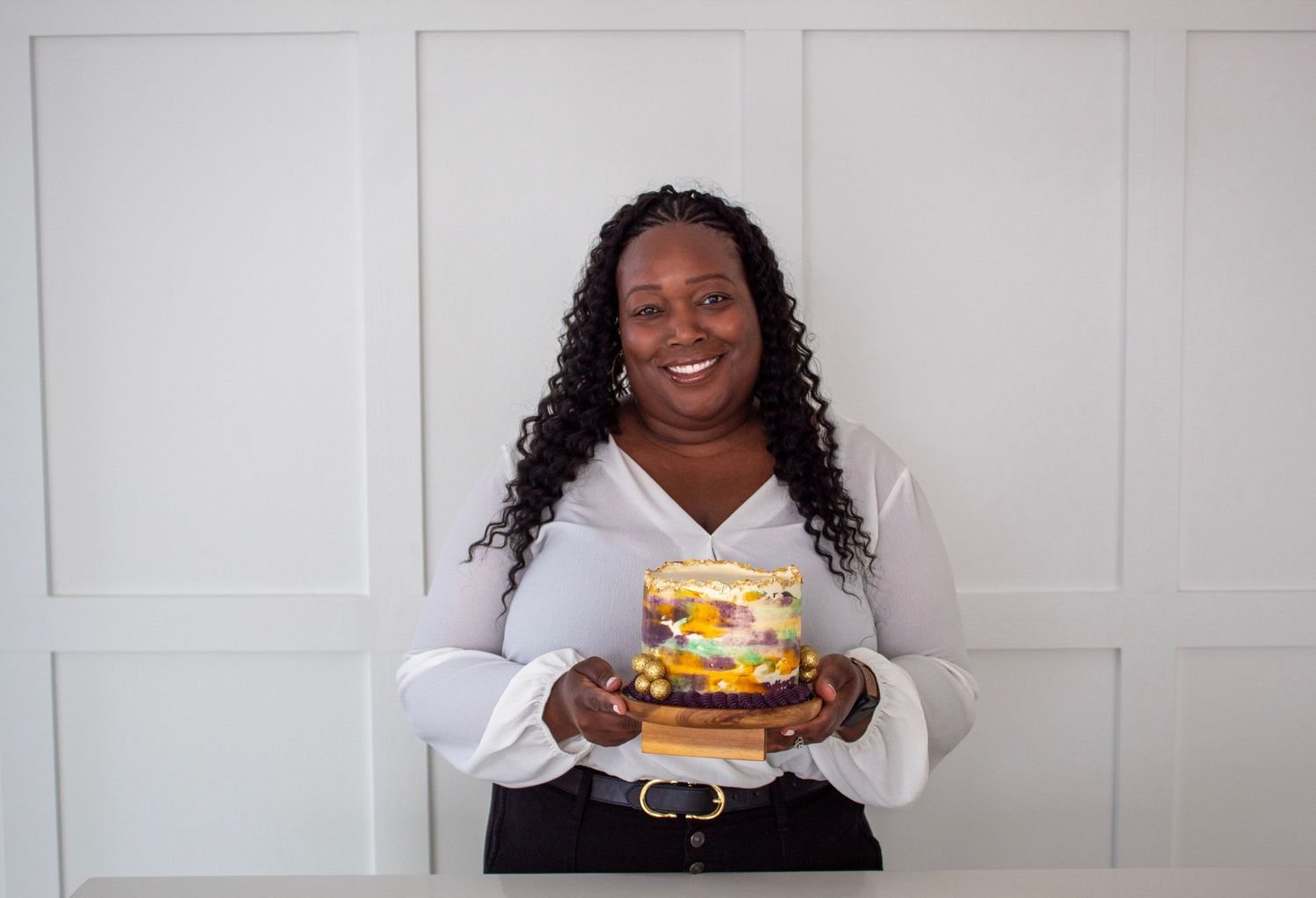 Smiling Black woman holds colorful decorated cake against white paneled wall background.