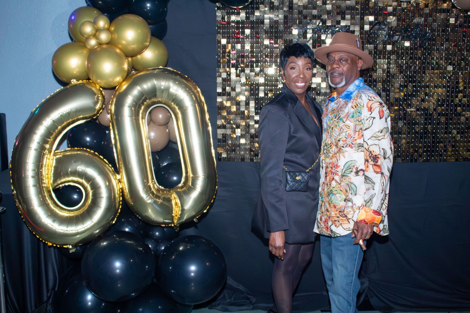 Couple posing together beside gold 60th birthday balloon arrangement at celebration party.