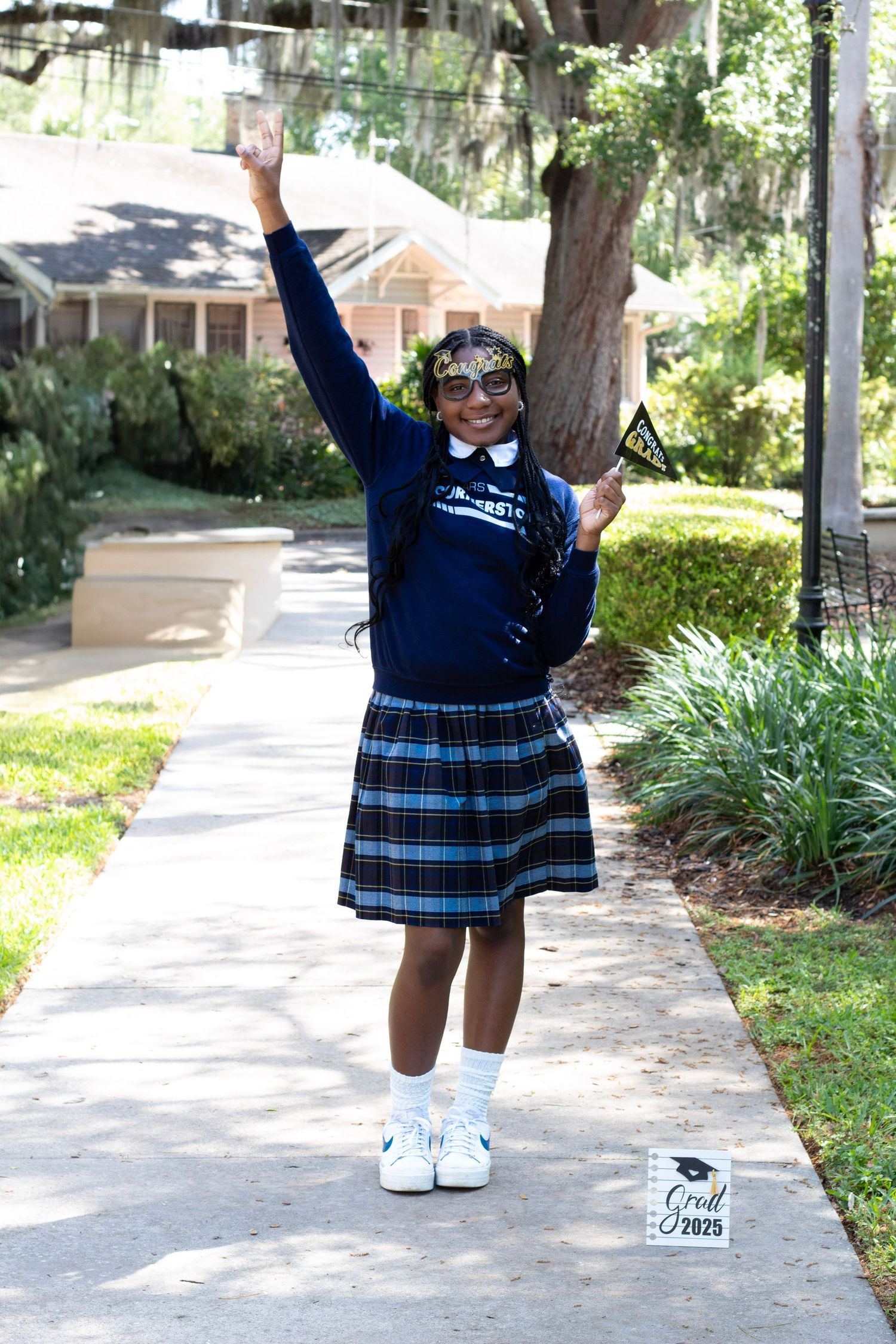 Excited graduate in navy school uniform raising hand on sidewalk with Grad 2025 sign nearby.