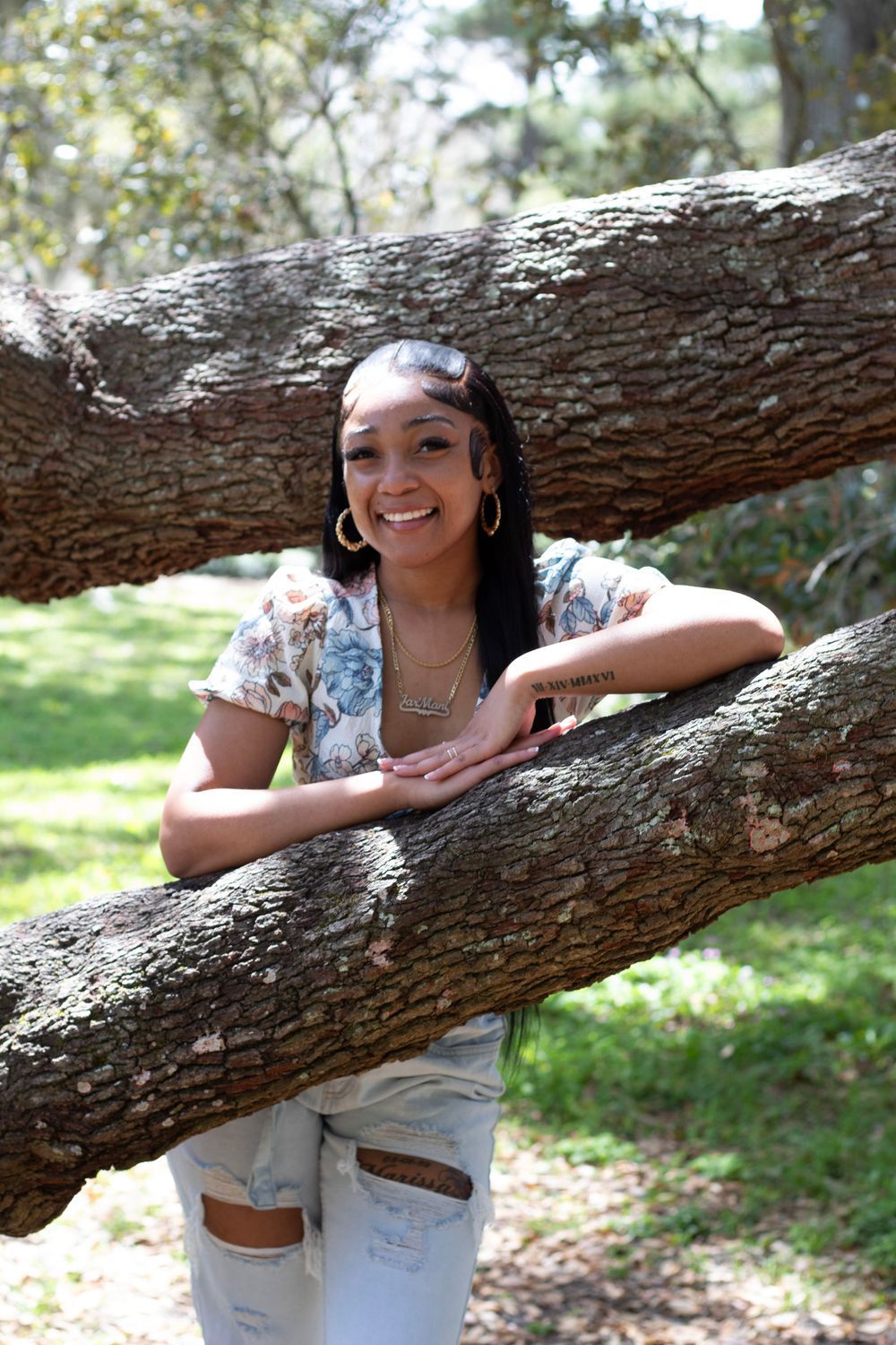 Smiling young woman leaning on large tree branches in a sunny park wearing floral top and ripped jeans.