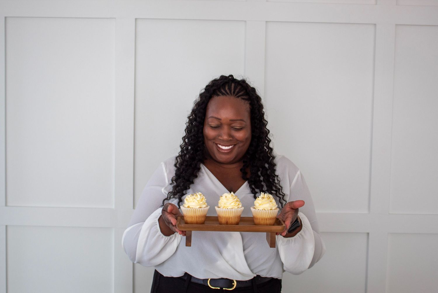 Smiling woman holding wooden tray of cupcakes with cream frosting against white paneled wall.