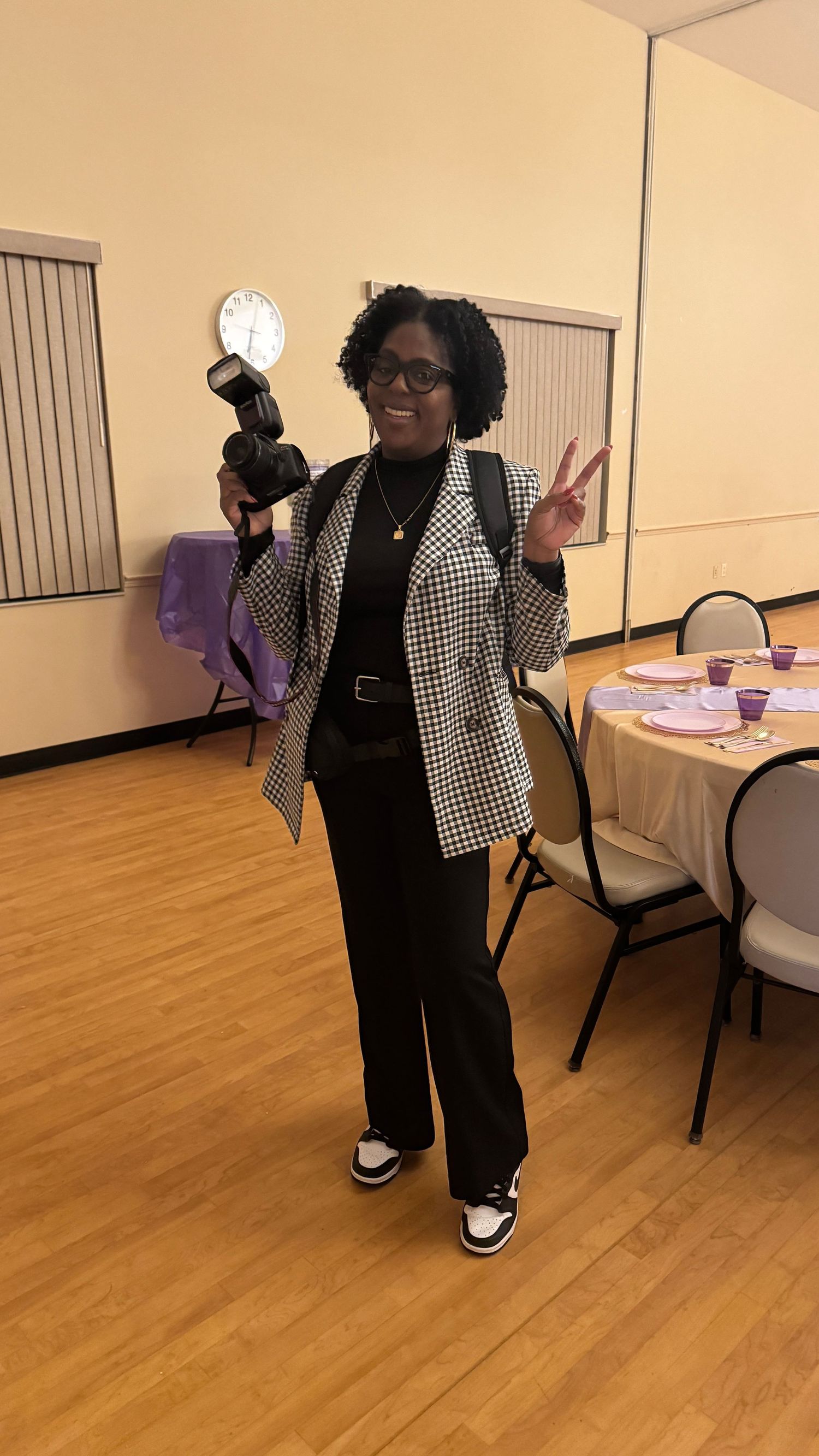 Smiling woman in houndstooth blazer holding camera, giving peace sign indoors.