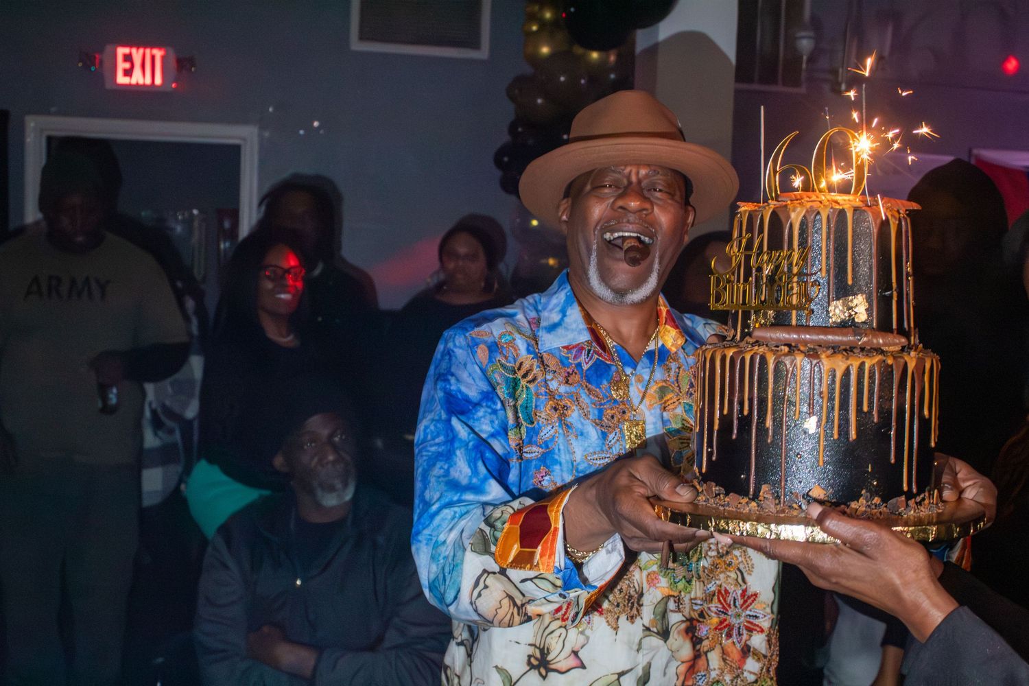 Man in floral shirt celebrating birthday holding sparkler-topped black and gold cake.