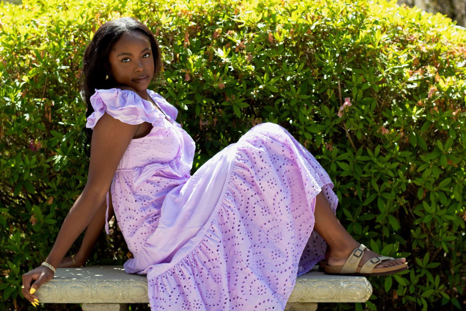 Young woman in lavender eyelet dress sitting gracefully on outdoor bench.