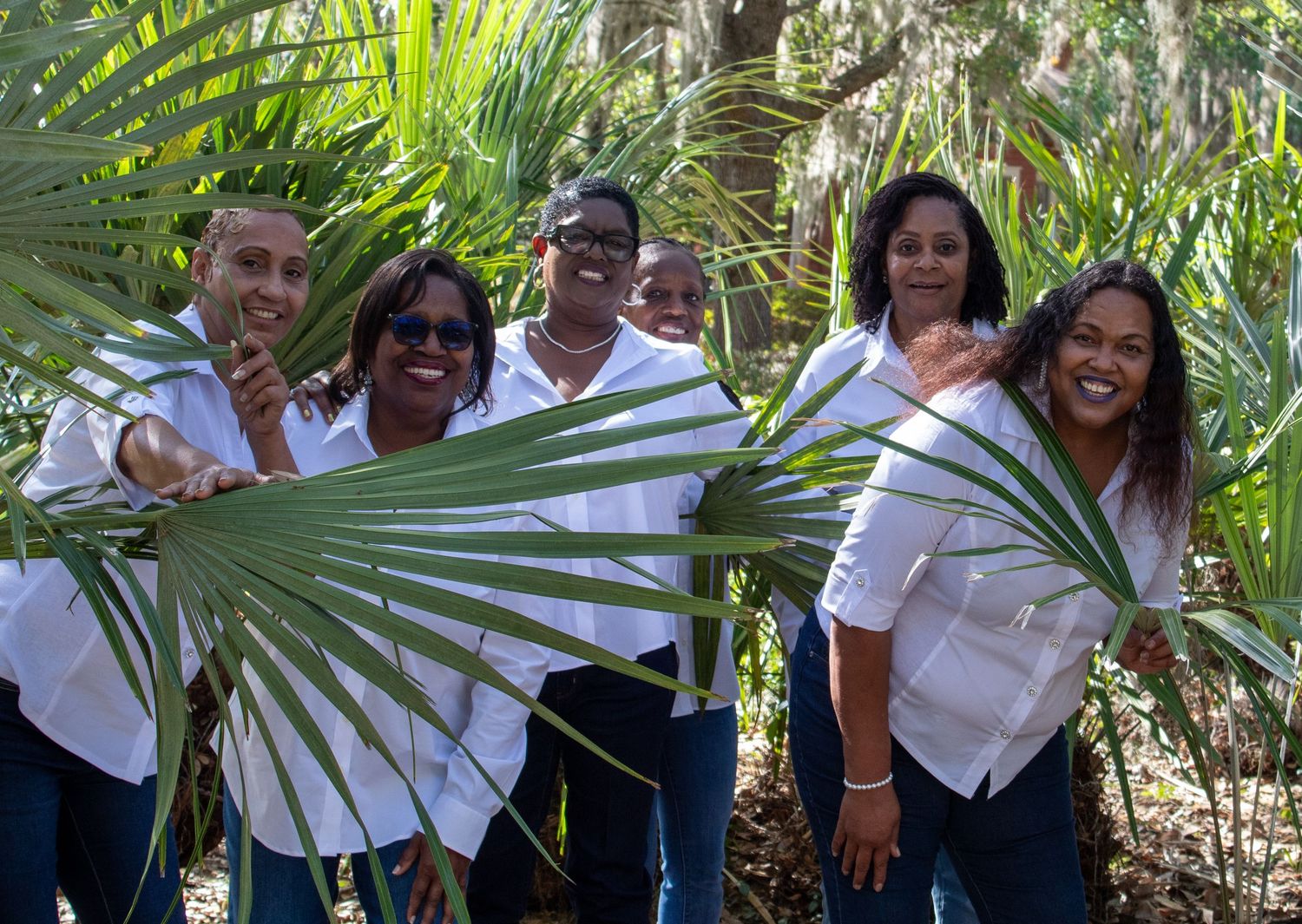 Group of five people in white shirts smiling behind large palm frond in lush tropical setting.