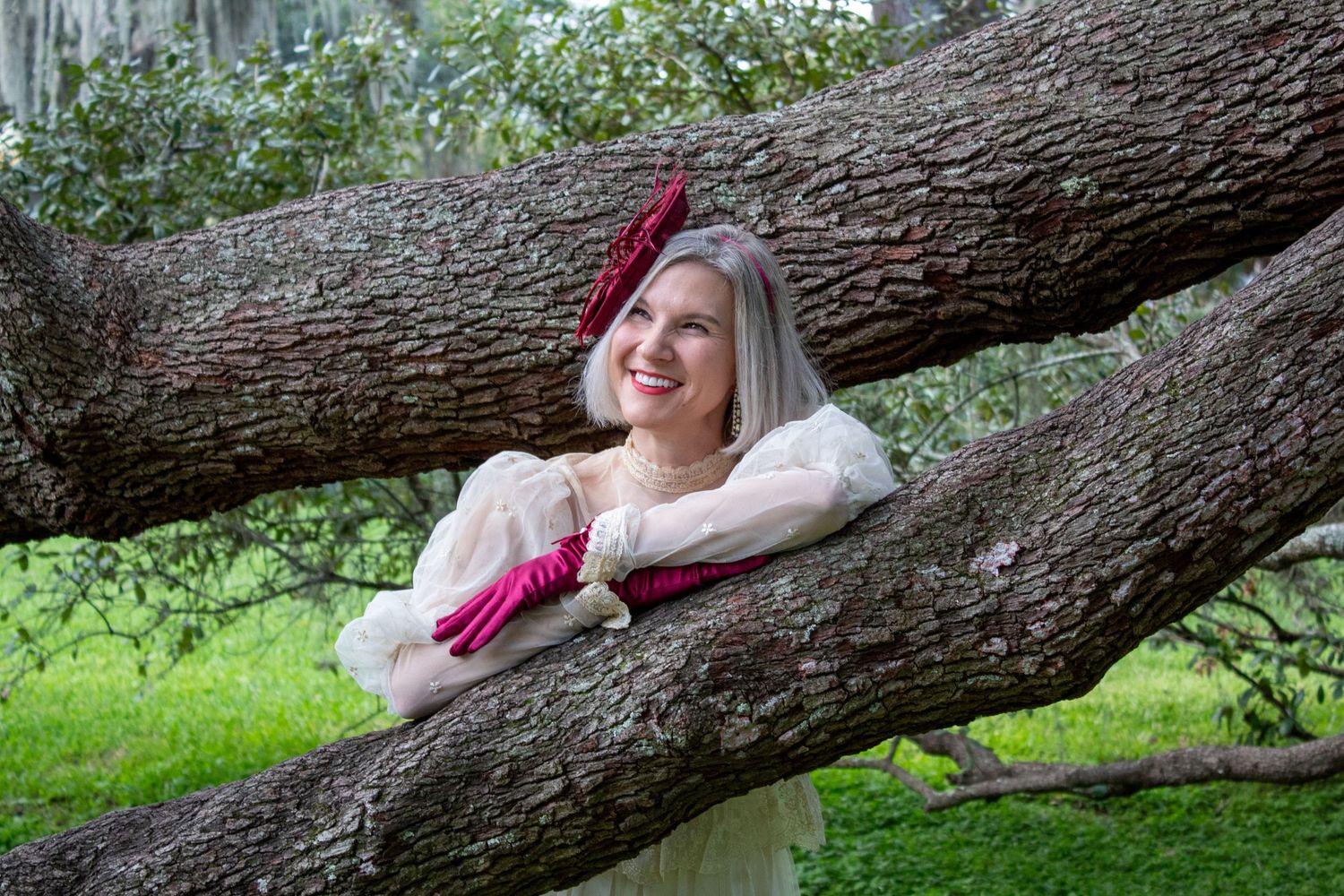 Elegant woman in vintage dress and red hat posing gracefully between large tree branches in a park.