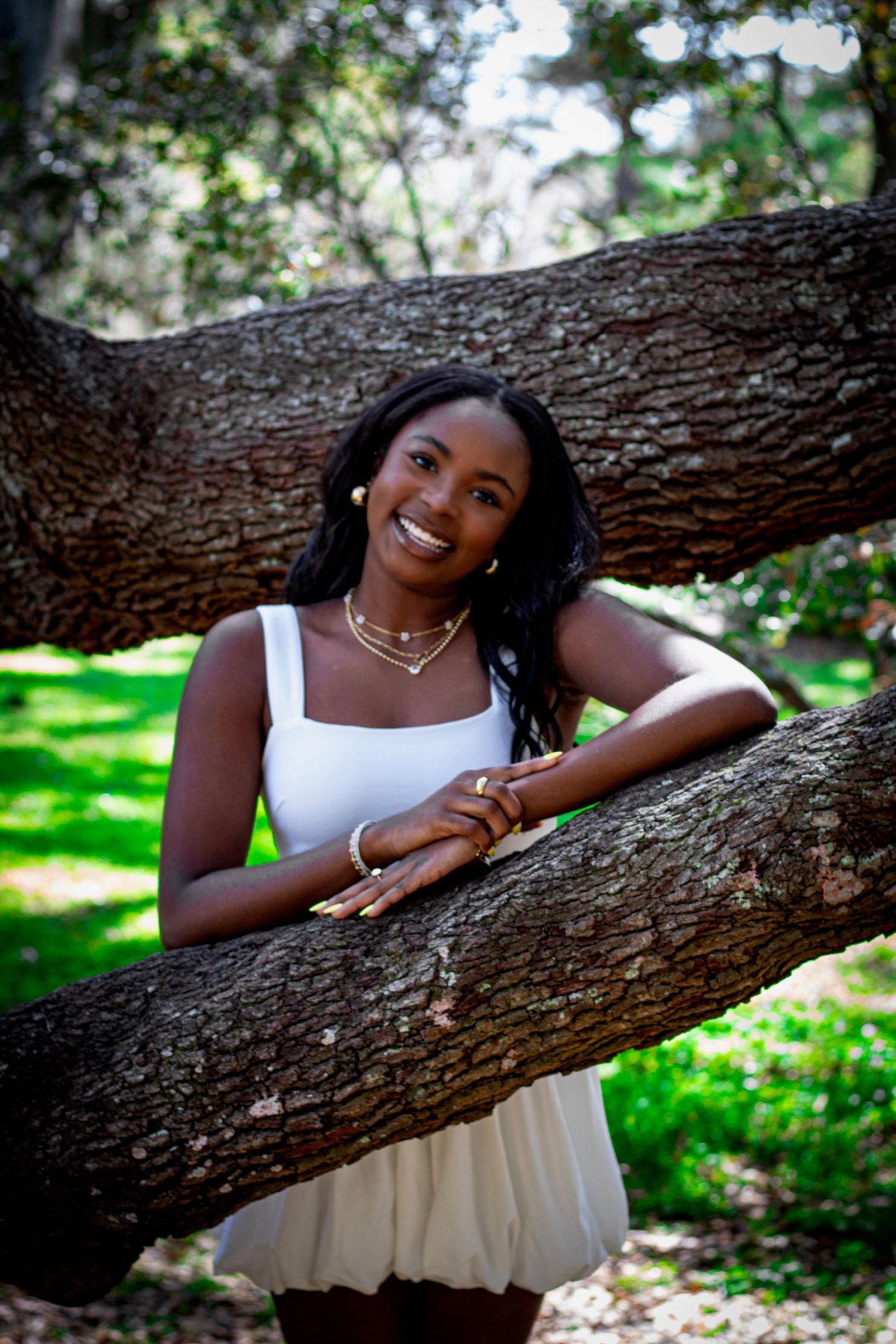 Young Black woman in white dress smiles leaning on tree branch in lush green park.