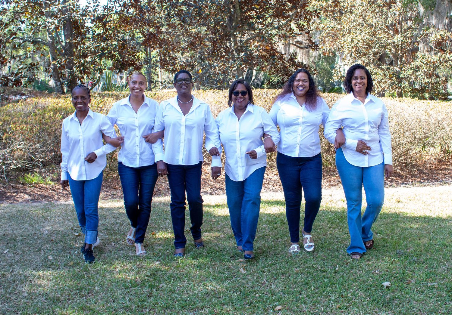 Six women in white shirts and jeans walking together and laughing in a sunny outdoor park.